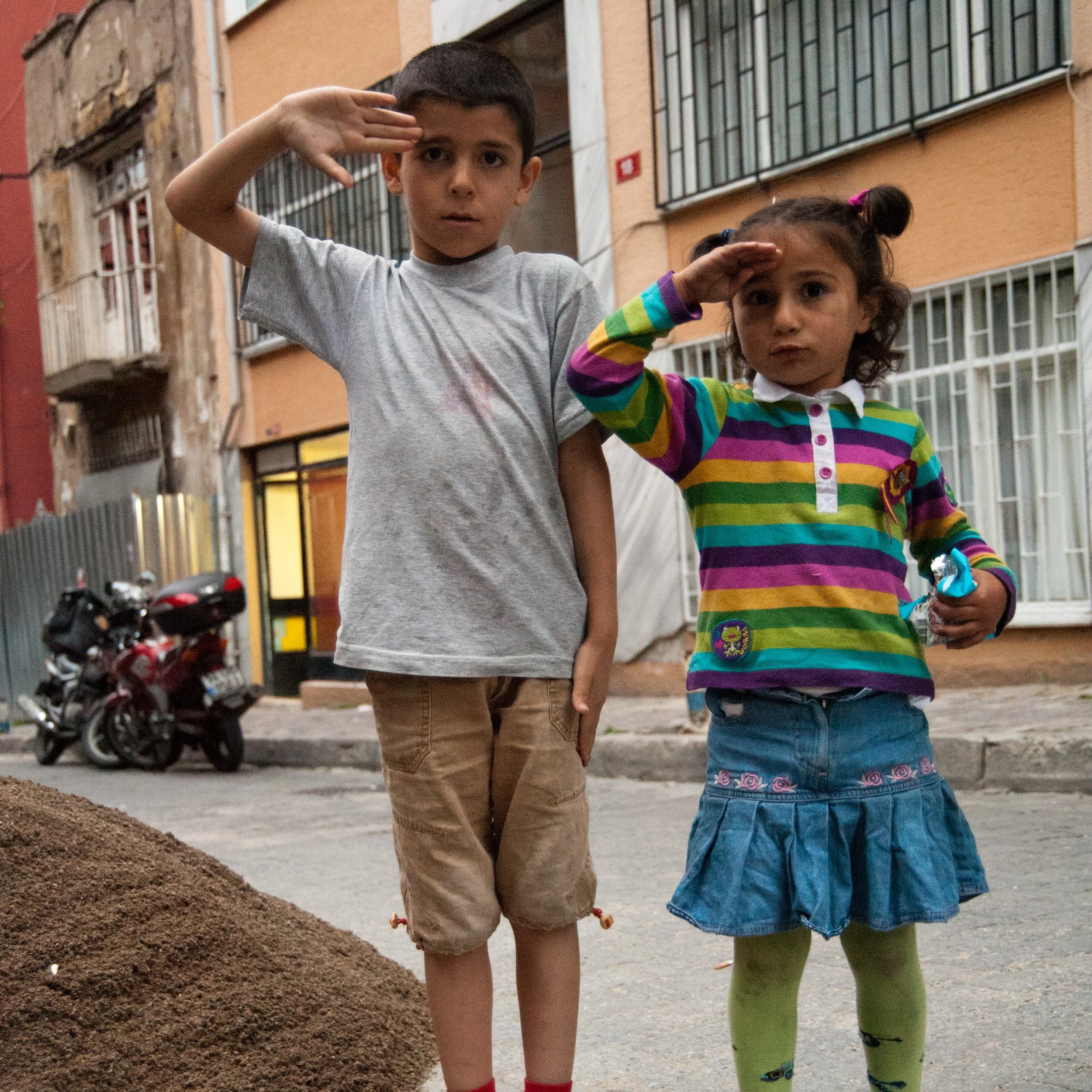 Two children, a boy and a girl, stand side by side on a city street, saluting with right hands raised to their foreheads. The boy wears a gray T-shirt and brown shorts, while the girl wears a colorful striped long-sleeve shirt, a blue denim skirt, an