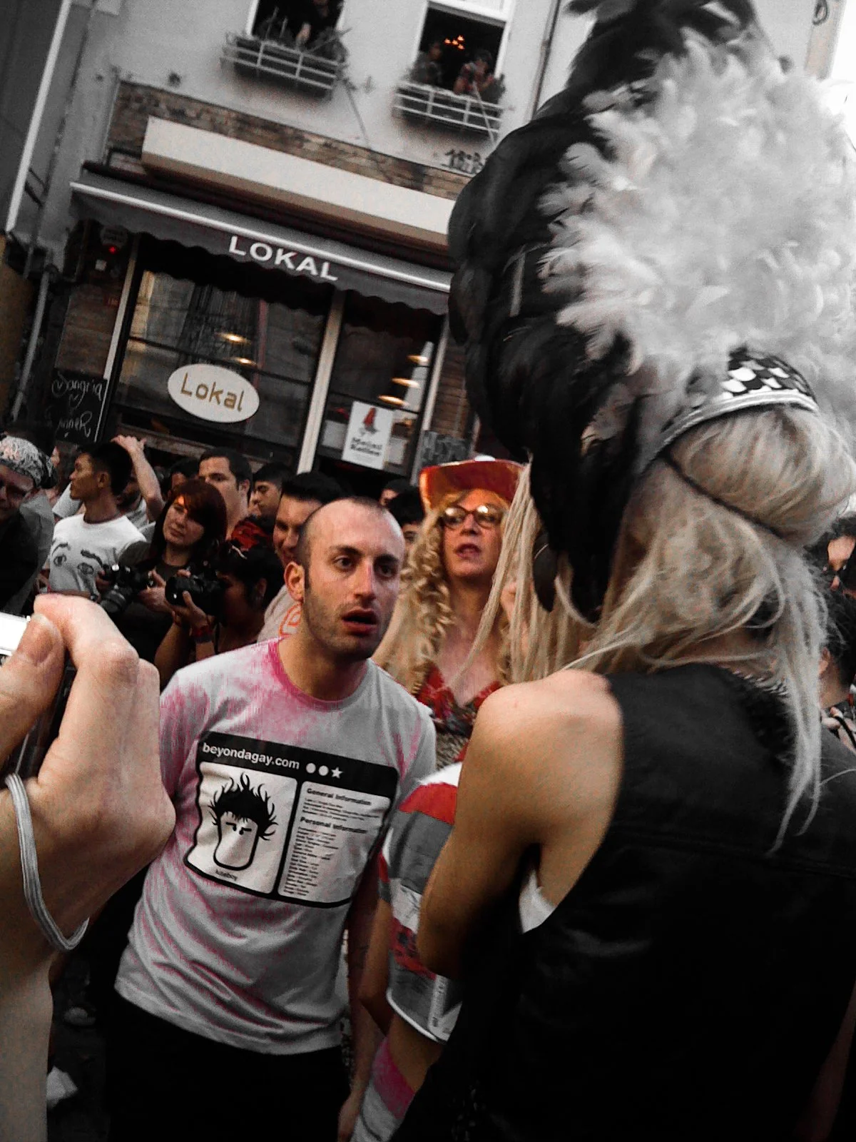 A crowd of people gathered outdoors at night, with some looking at a person dressed in a costume with a furry mask and long hair. The setting appears to be a city street.