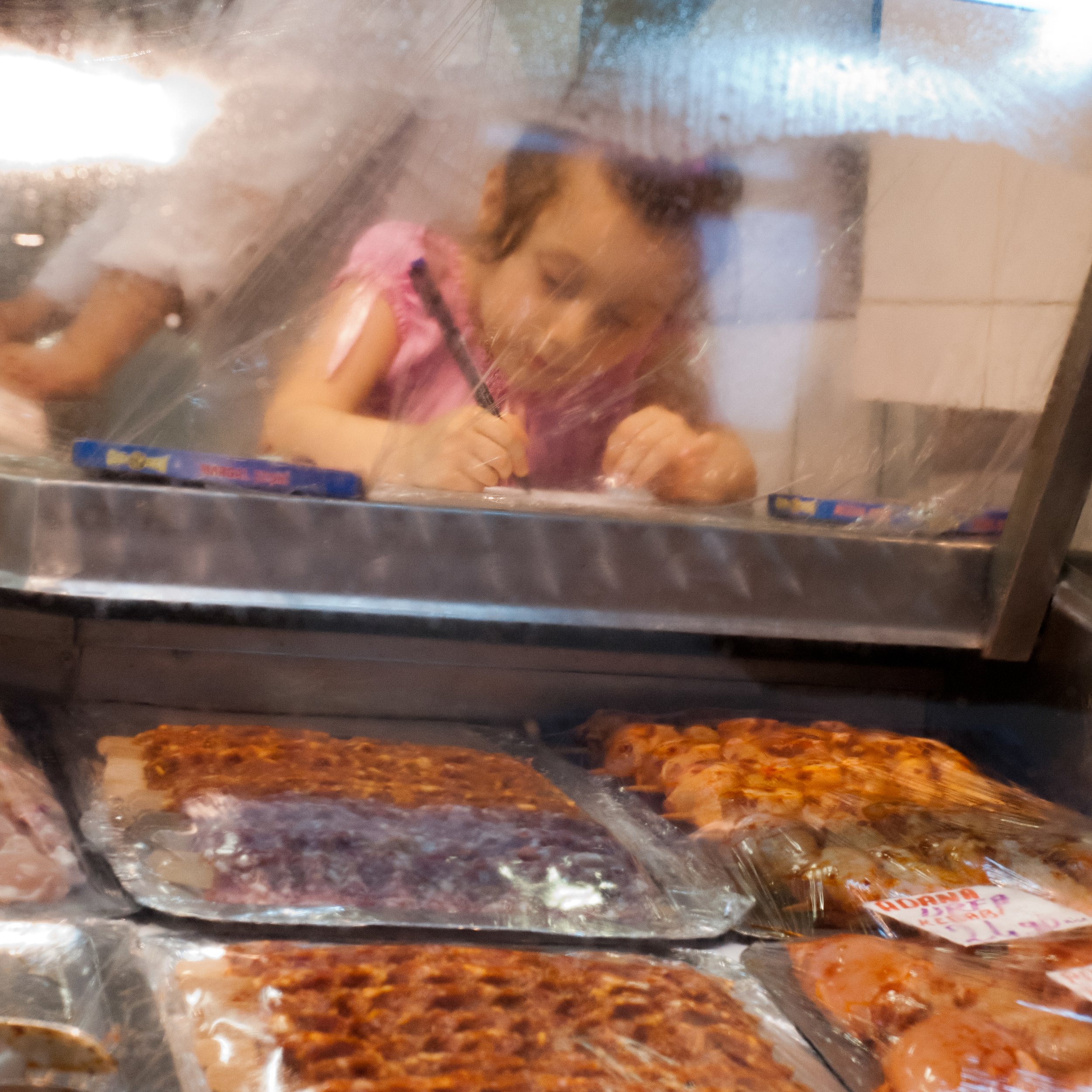 A young girl is writing with a marker inside a plastic-covered food display case at a pizzeria, with pizzas and packaged food items visible in the case below.