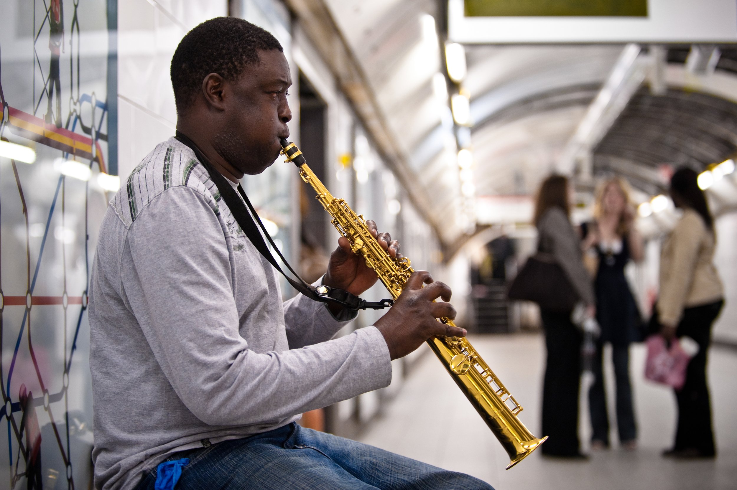 A man playing a saxophone on a subway platform, with three women talking in the background.