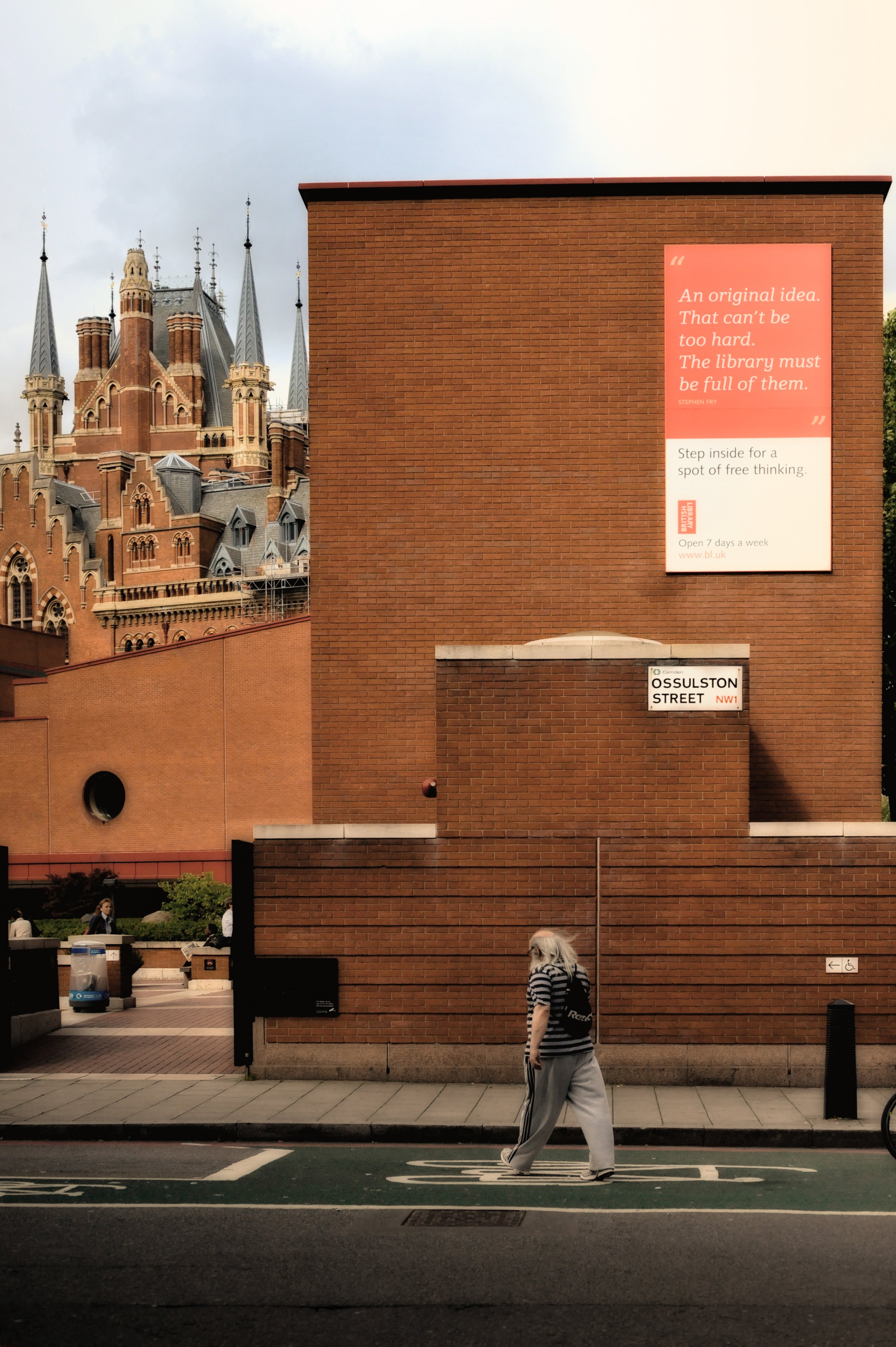A person walking on a city street with a brick building and a historic castle-like building in the background; the brick building has a sign that says 'Ossulston Street'.