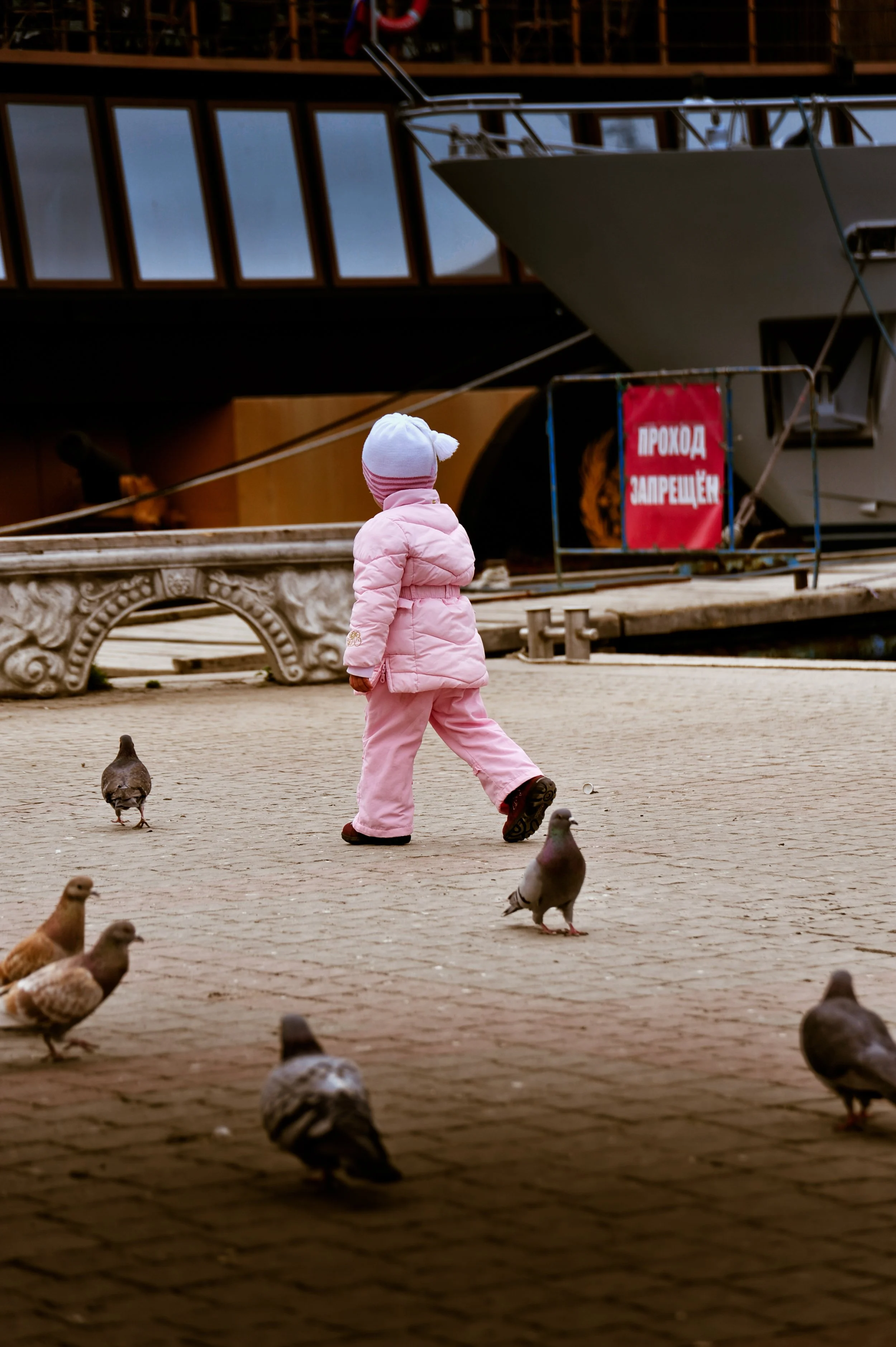 A young girl dressed in pink clothing and a white hat walking among pigeons on a paved waterfront area, with a ship and a red sign in the background.