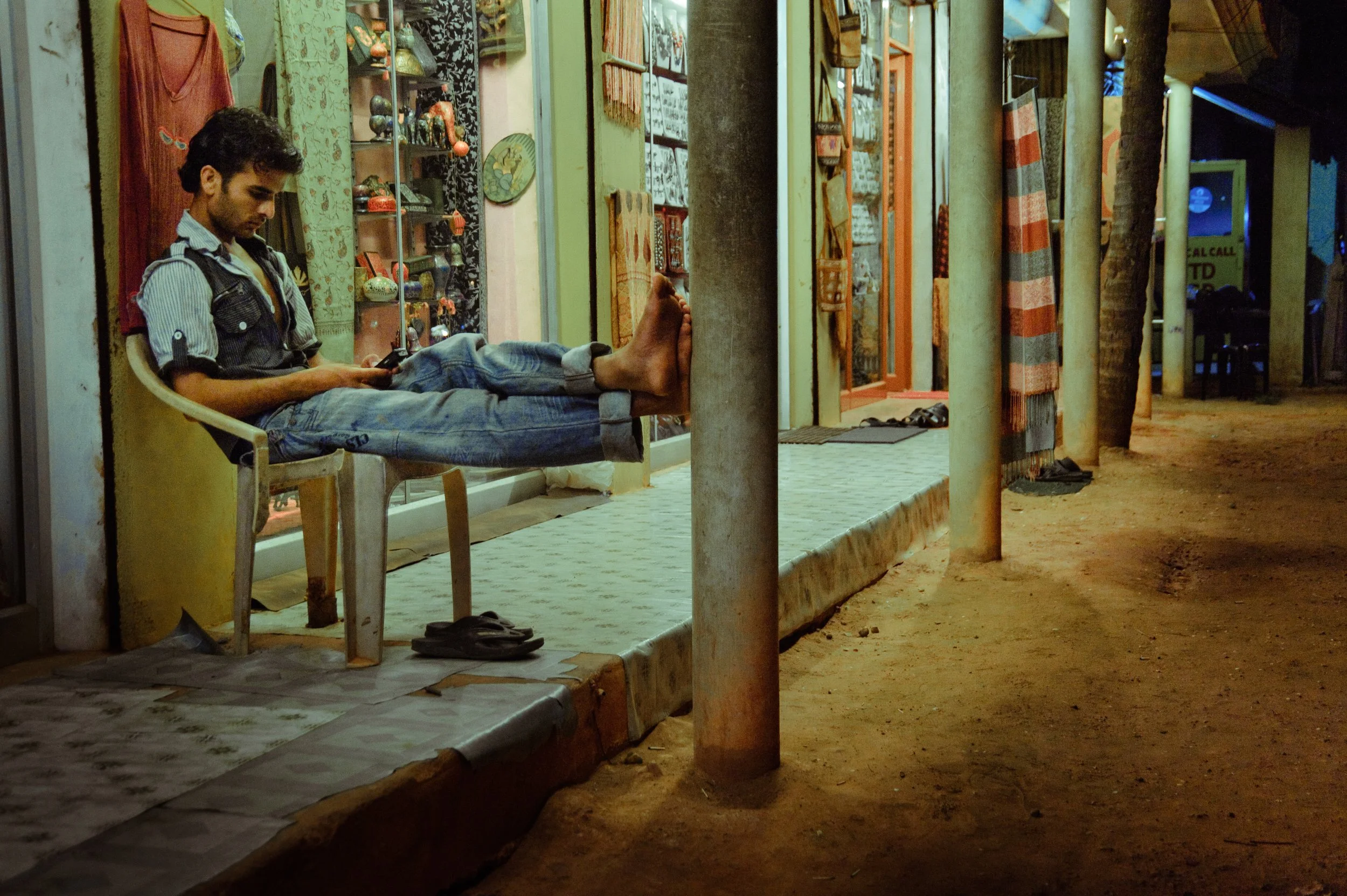 A young man with a beard and dark curly hair is sitting on a plastic chair outside a shop at night. He is wearing a striped shirt with rolled-up sleeves, a black vest, and jeans. His legs are stretched out and resting on a nearby pole as he looks at 