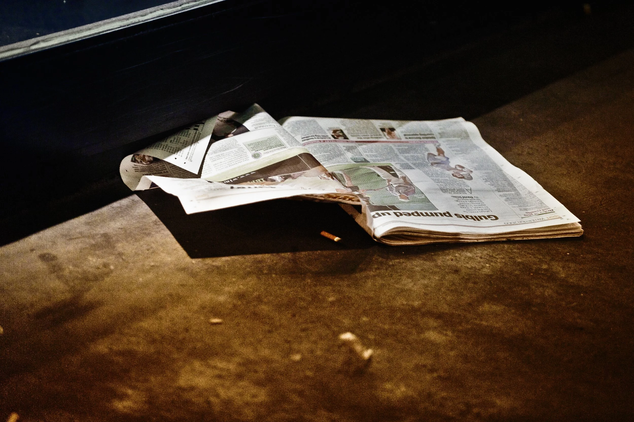 A crumpled newspaper lying on a concrete ground under a car, with part of the vehicle visible at the top of the image.