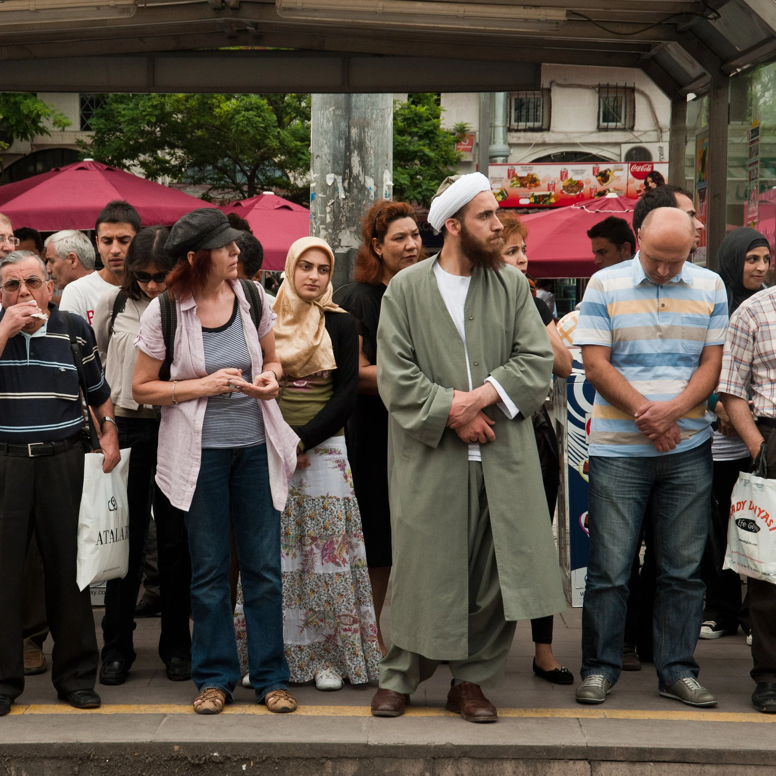 A diverse group of people standing at a bus stop or waiting area, some with heads bowed or eyes closed, outdoors with trees, umbrellas, and food stands in the background.