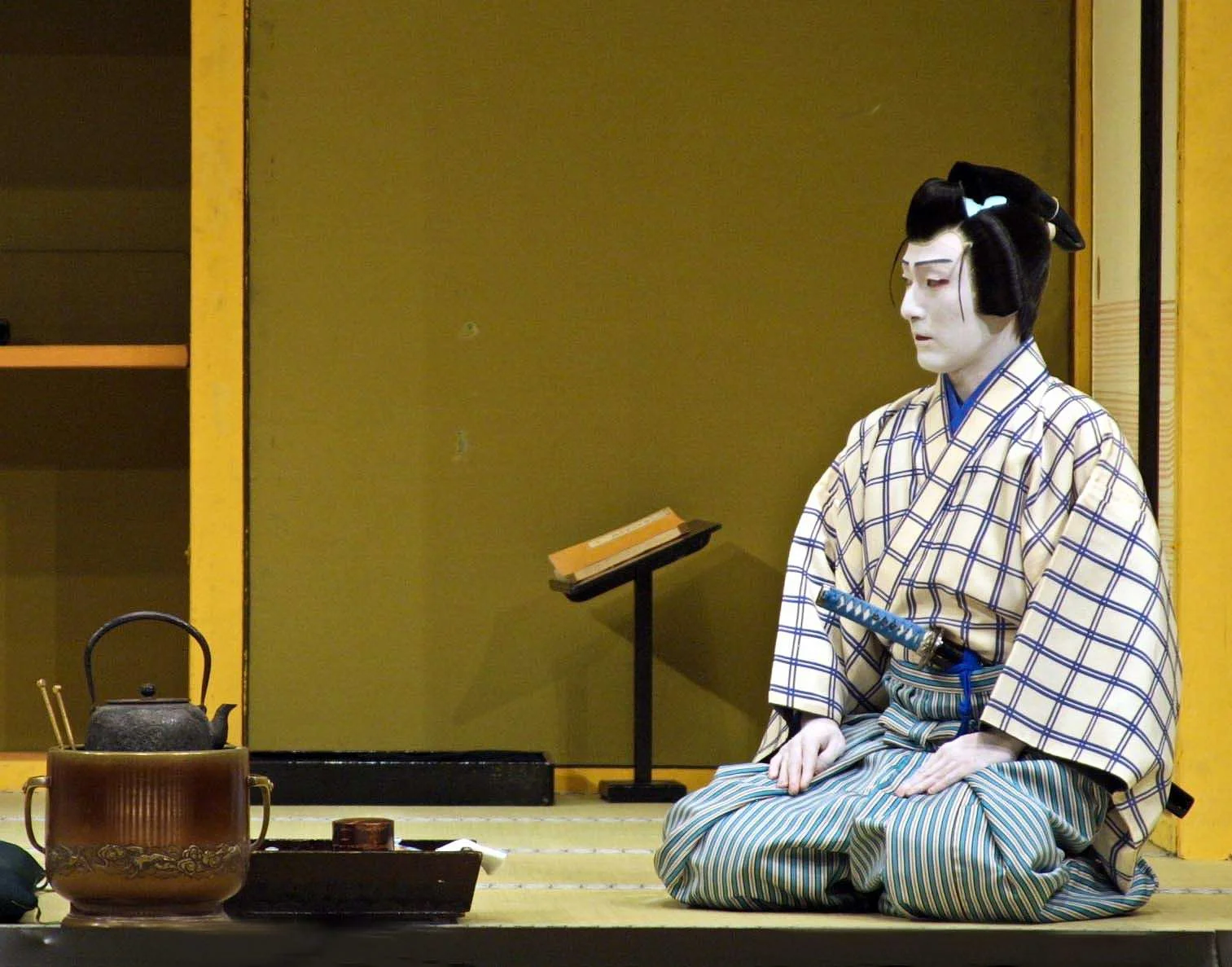 A traditional Japanese puppeteer in costume and makeup performing a Bunraku puppet show on stage, with puppets and props visible in front.