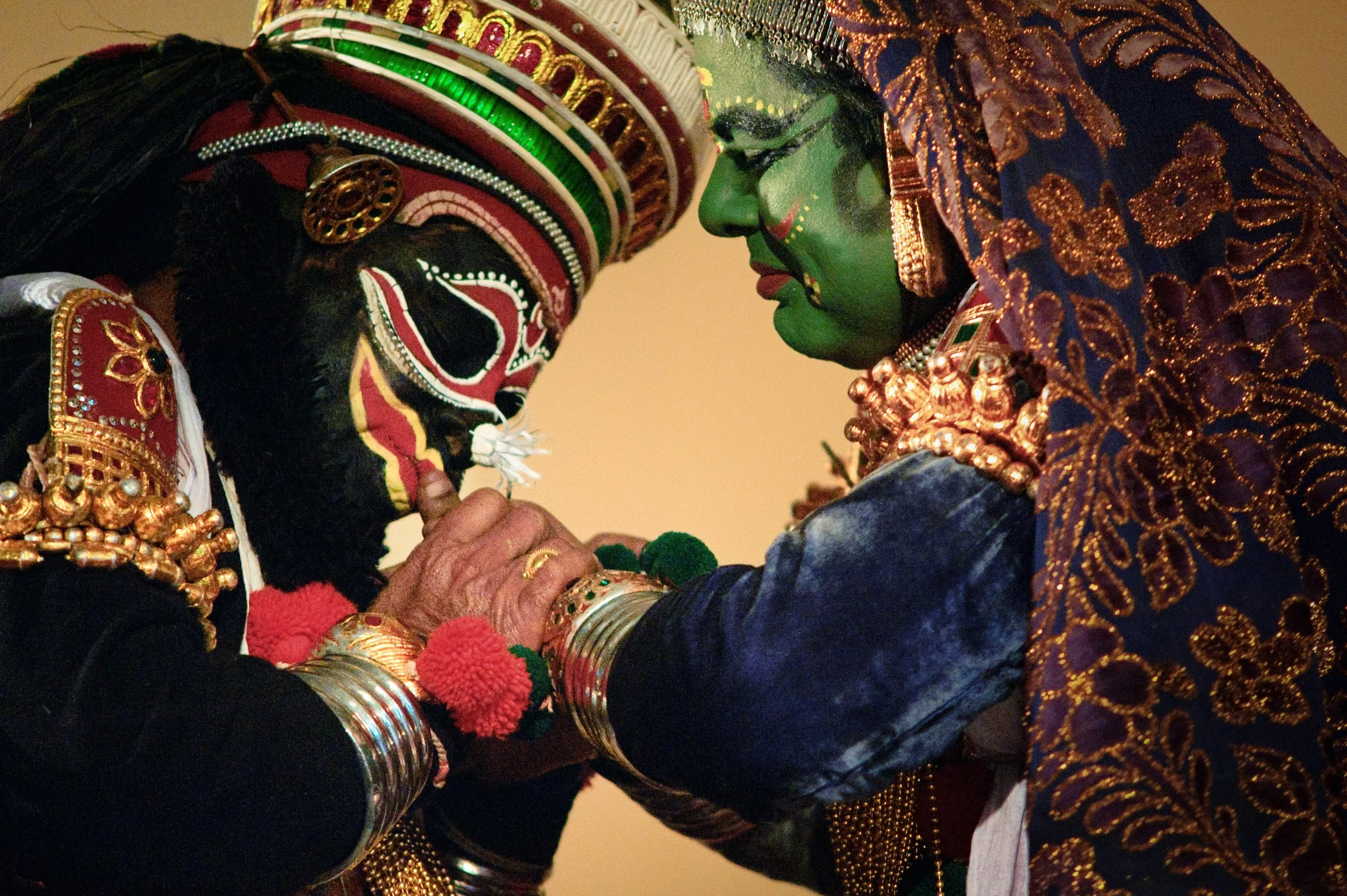 Two individuals dressed in traditional Indian attire, with one person painted as Lord Shiva and the other as Goddess Kali, touching foreheads in a ritual.