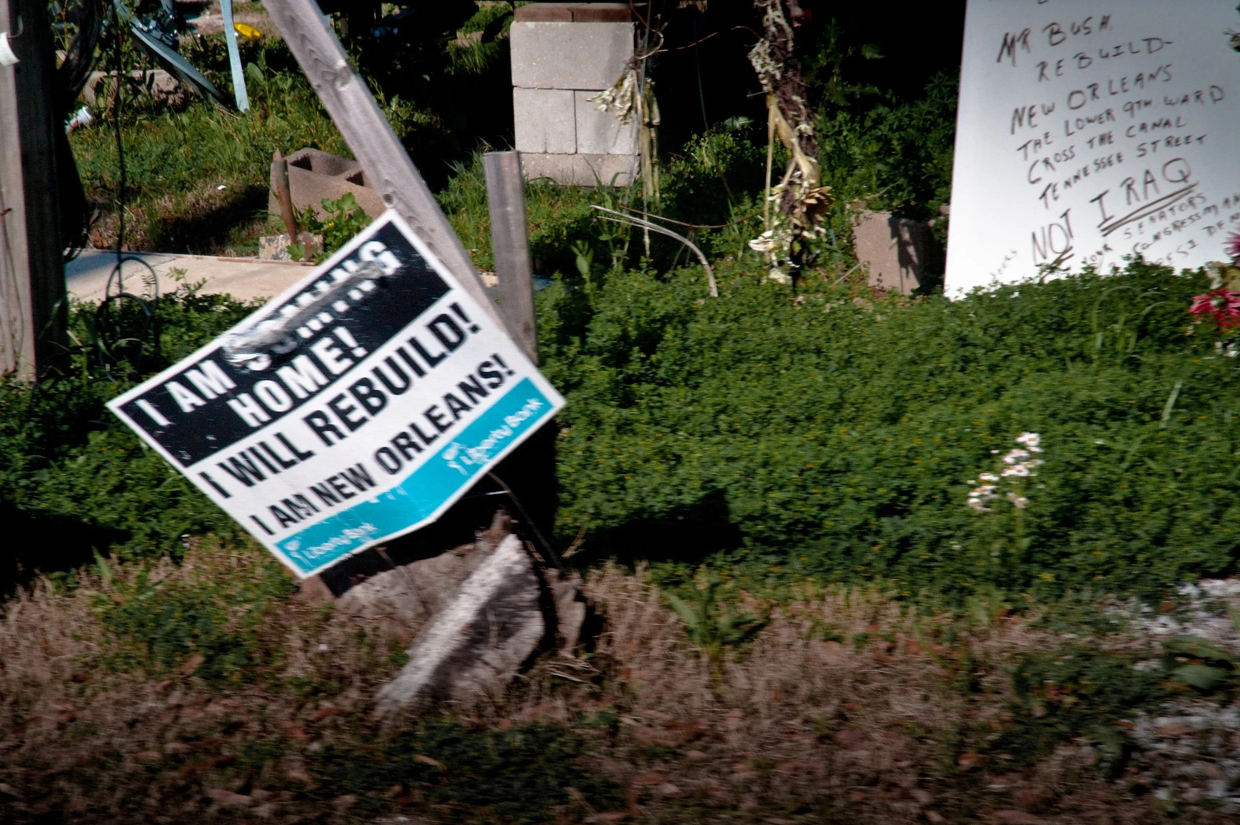 Signs in a garden area supporting the rebuilding of New Orleans after a flood, with one sign reading "I AM SEEING! HOME! I WILL REBUILD! I AM NEW ORLEANS!" and a handwritten sign in the background with additional messages about New Orleans.