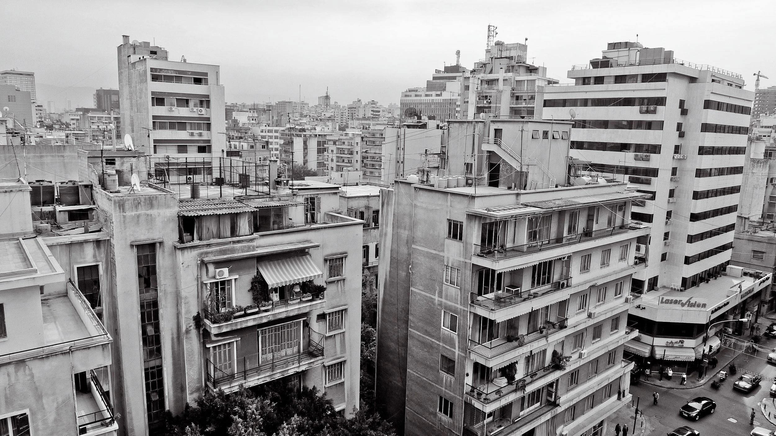 Black and white photo of multiple tall apartment buildings with balconies and rooftops, cityscape background, streets with cars and pedestrians at street level.