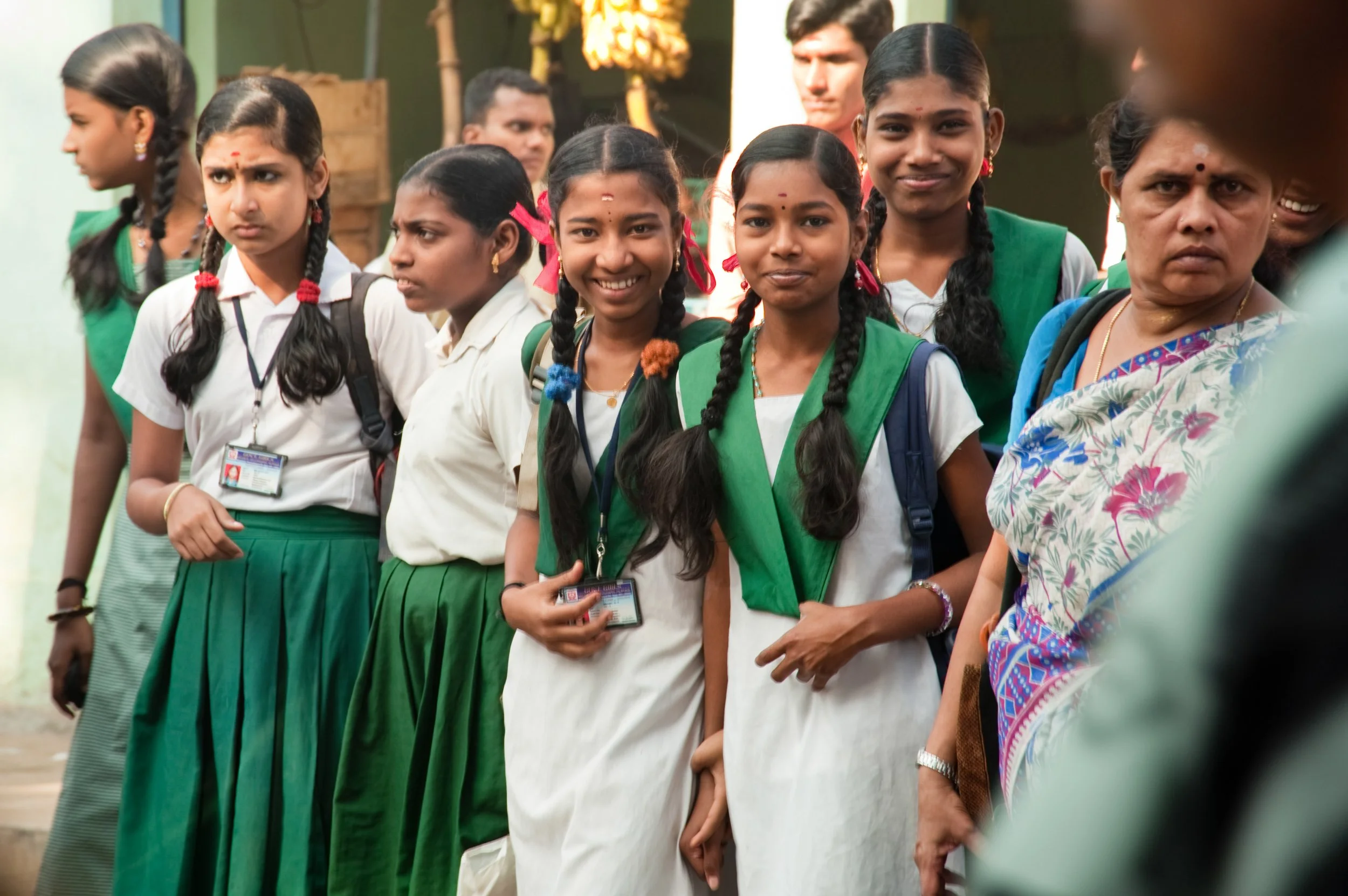 Group of schoolgirls in uniform standing in line outdoors, some smiling and others looking serious, with a teacher or adult woman partially visible on the right.