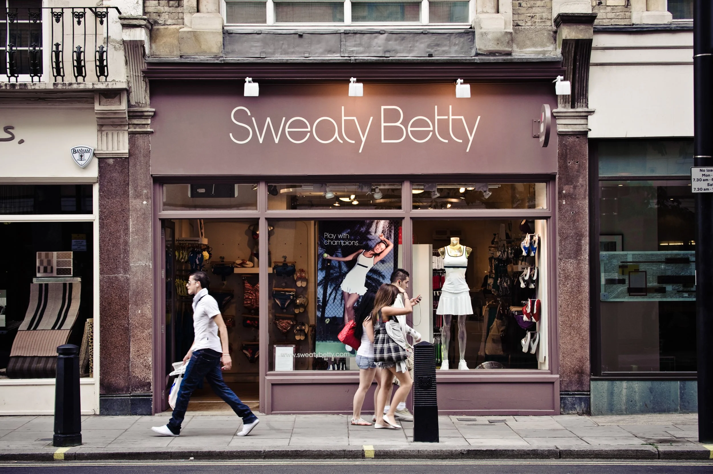  storefront with purple facade labeled 'Sweaty Betty' featuring a large poster of a woman in tennis attire and a mannequin in a white dress, with three pedestrians walking on the sidewalk in front.