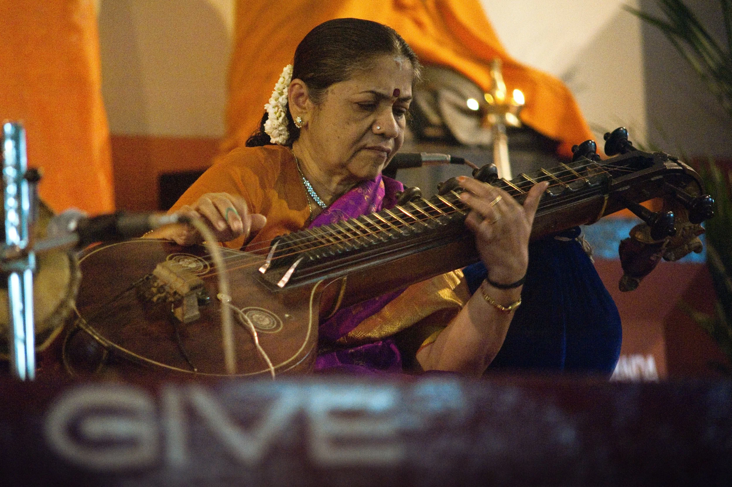 An elderly woman with traditional Indian attire playing a sitar in a performance setting.