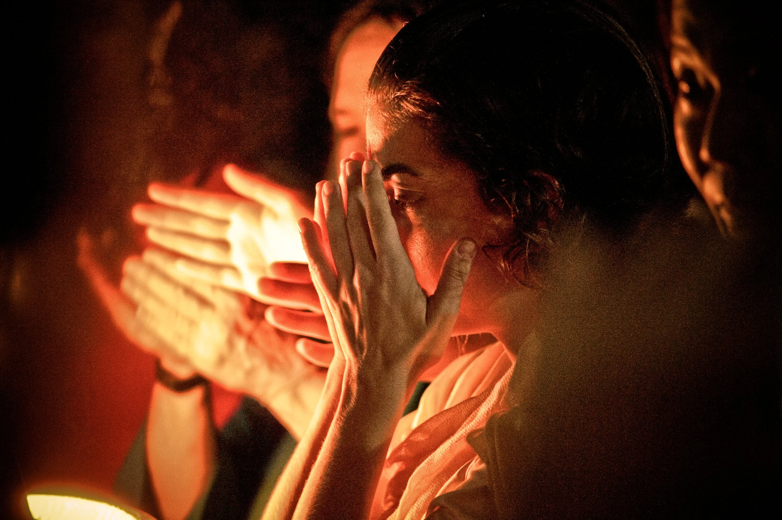 A woman with short hair covering her mouth with both hands in a dark setting, illuminated by a warm orange glow.