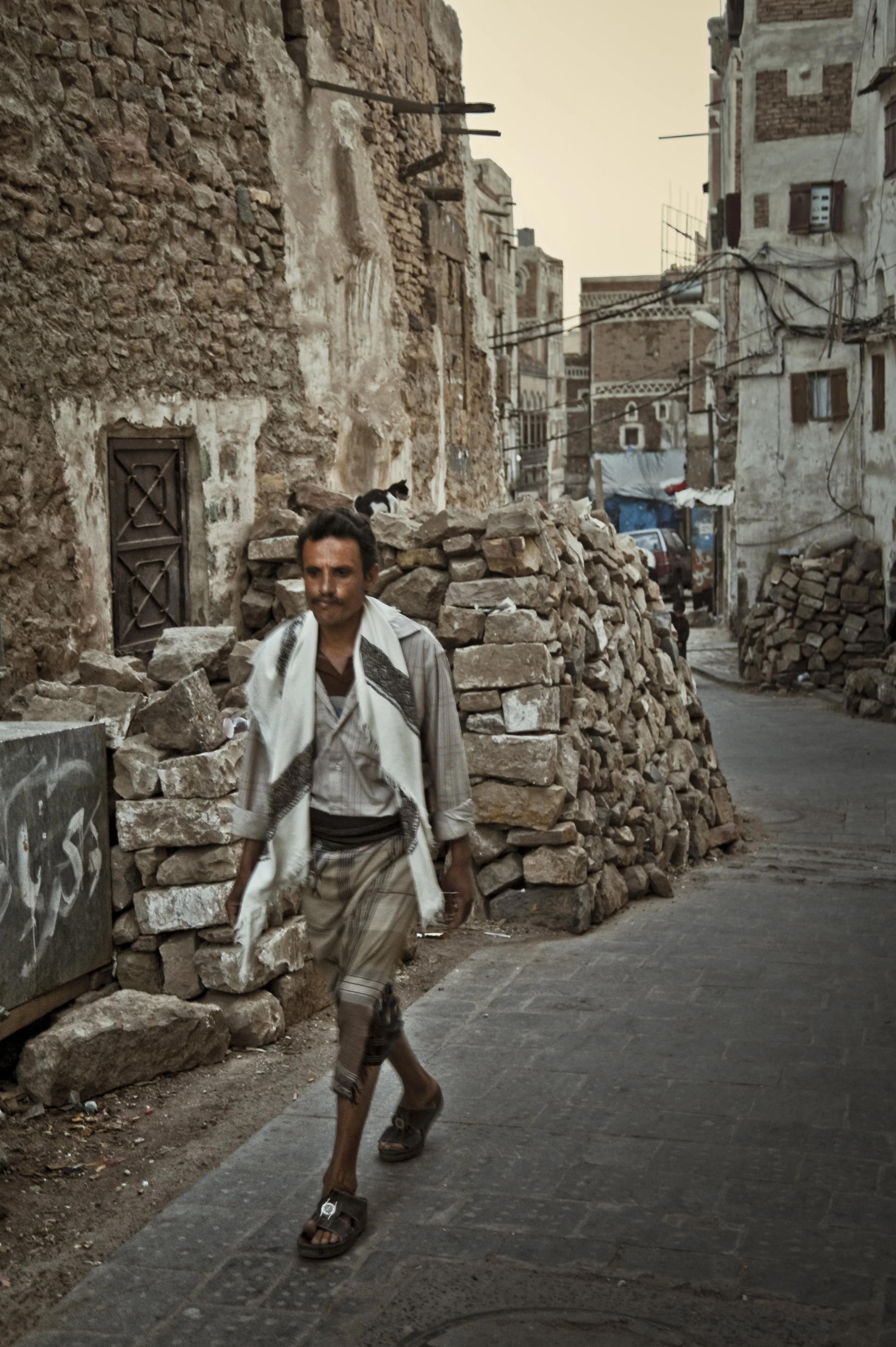 A man walking down a narrow alleyway with stone and brick piles on either side. The alley is surrounded by tall, old, weathered buildings with exposed wiring and a few cats on top of the stones. The scene appears to be in a historic or older part of 