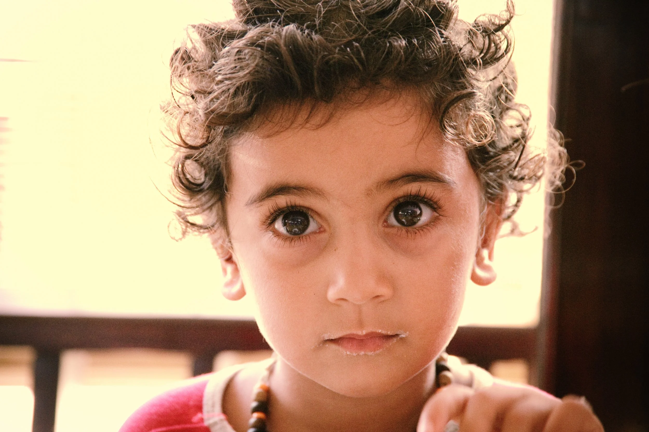 Close-up of a young child with curly hair and big, dark eyes, wearing a red shirt and a beaded necklace, looking directly at the camera with a curious expression.