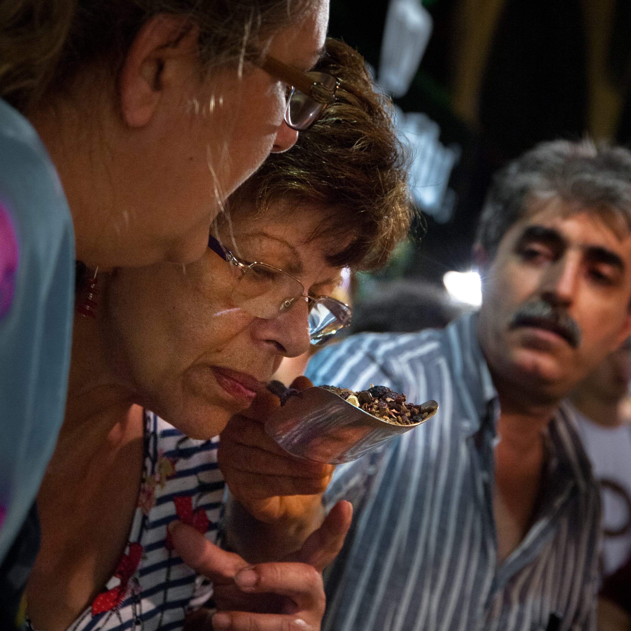 Three people, two women and one man, gathered closely at a table; the woman in the middle is tasting something from a spoon held by the woman on the left, with the man watching. The focus is on their faces and the spoon.