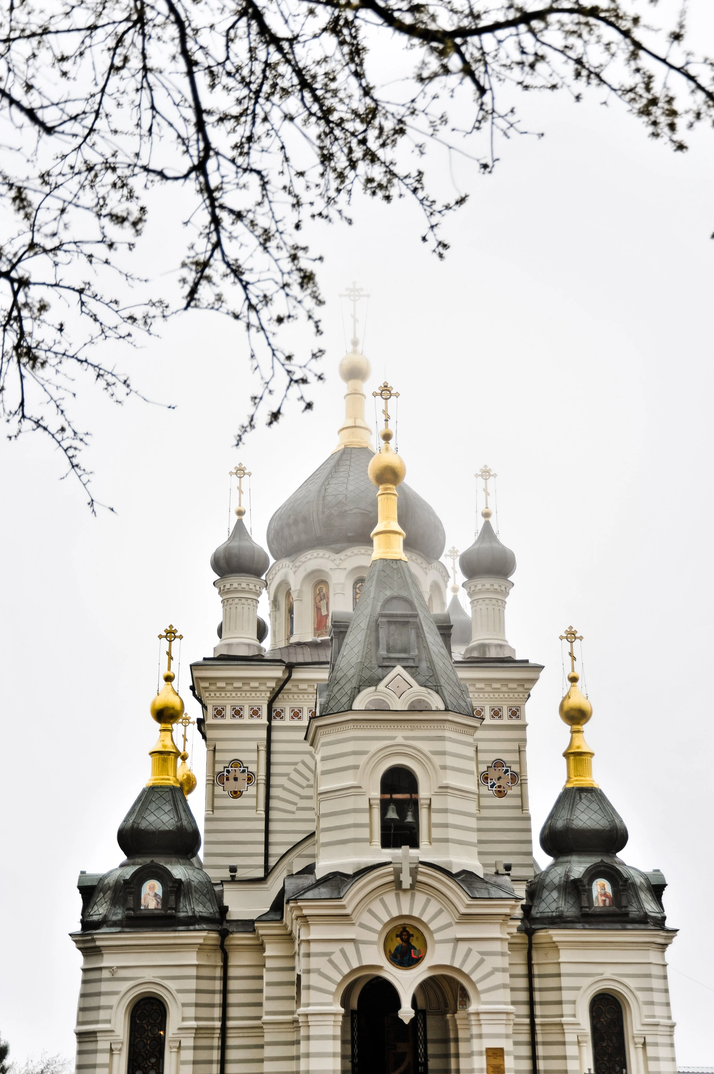 A church with multiple onion domes topped with crosses, featuring ornate architectural details, set against a cloudy sky with tree branches at the top.