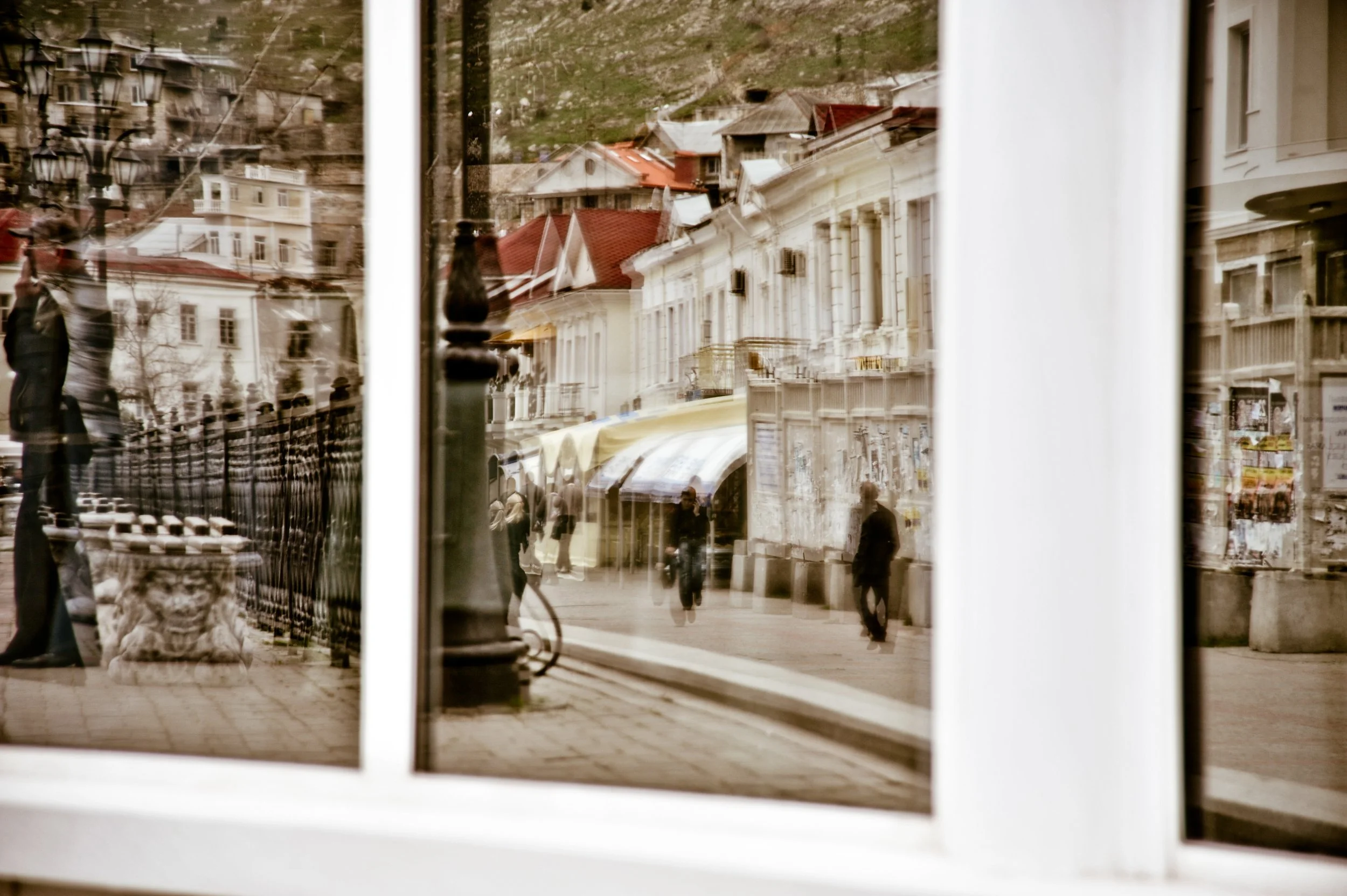 Reflected street scene in a window, showing pedestrians, buildings with red roofs, and a sidewalk with shops and street lamps.