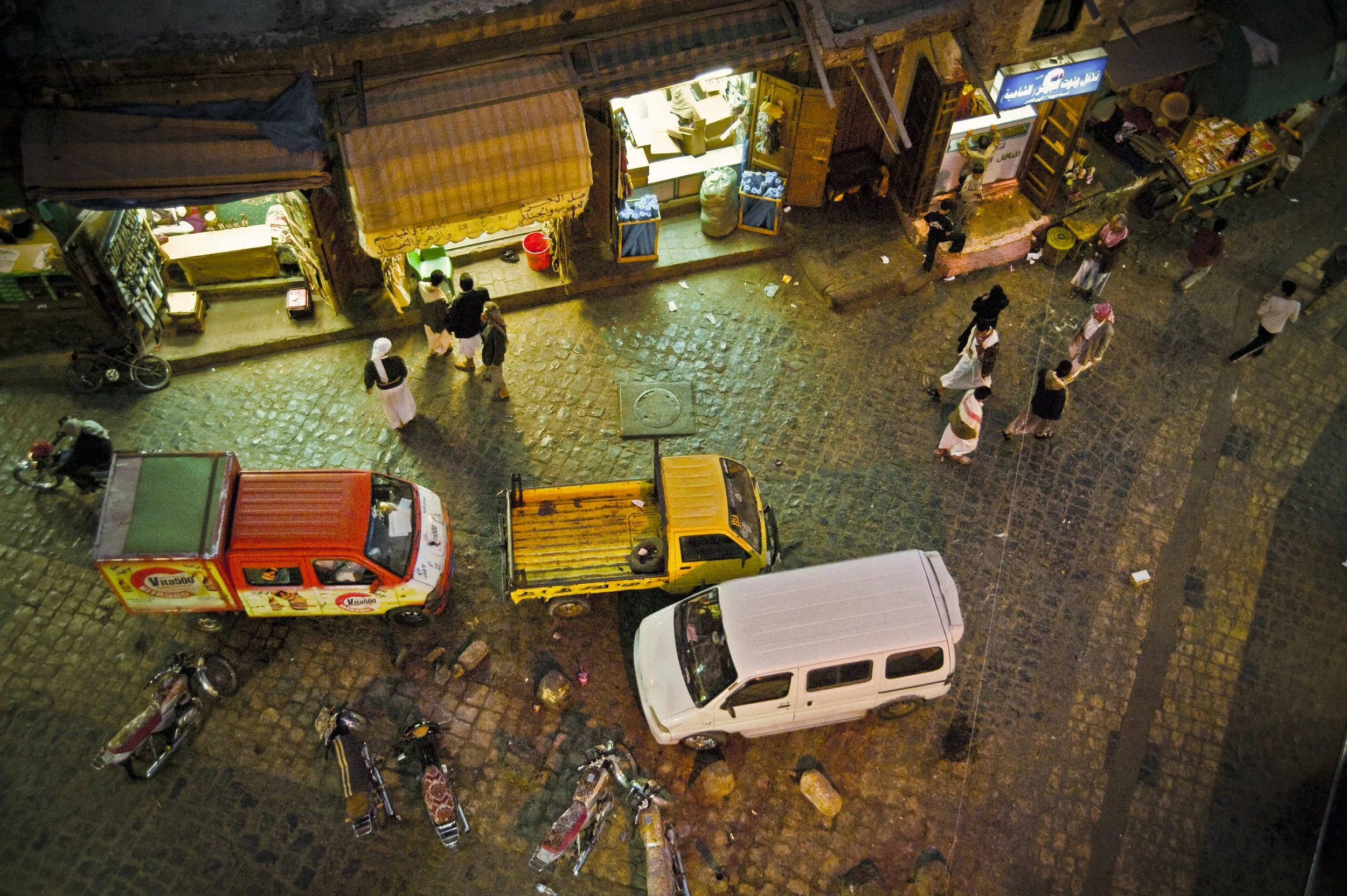 A nighttime street scene viewed from above featuring storefronts and pedestrians walking on a cobblestone road. There are parked motorcycles and cars, including a white van, a yellow truck, and a red delivery truck, with some street debris visible.