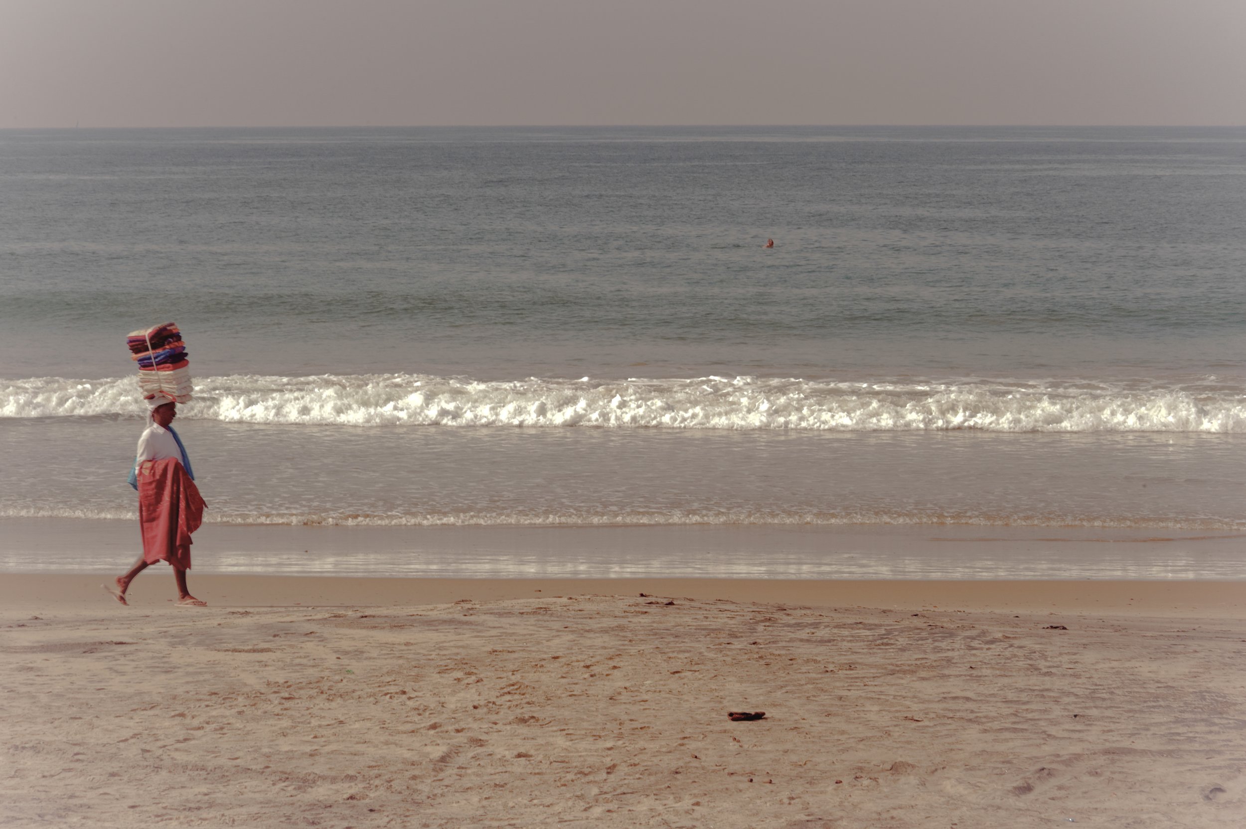 A woman walking along the sandy beach carrying a stack of colorful towels on her head, with an ocean and small waves in the background.
