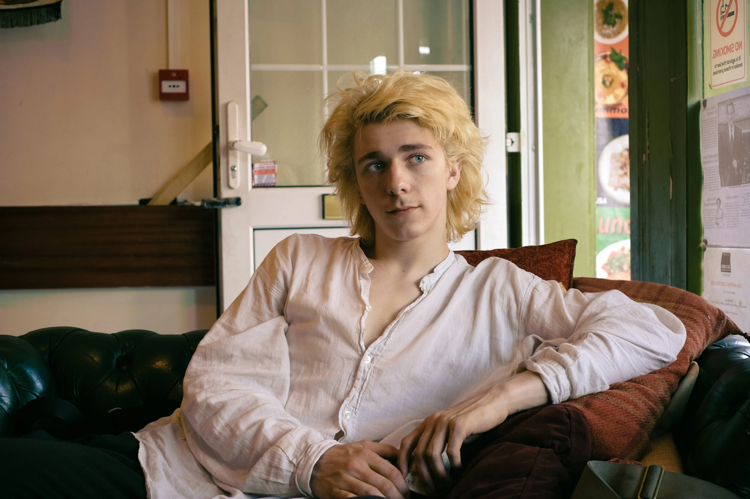 A young man with long, curly blonde hair sitting on a black leather couch inside a cafe or diner. He is wearing a cream-colored, slightly wrinkled button-up shirt and looking off to the side. Behind him, a door with glass panels and food menu posters