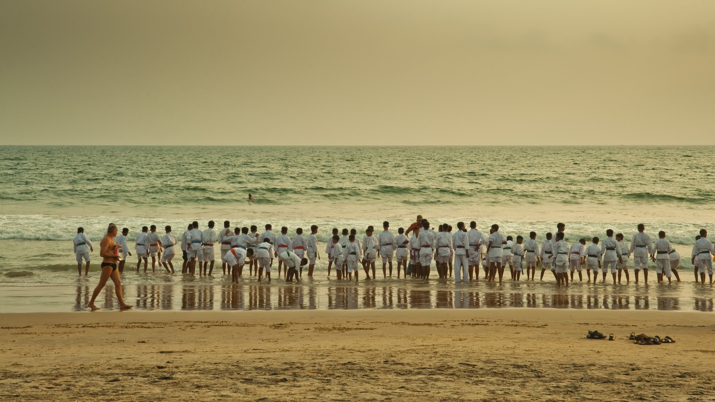 A group of martial arts students in white gis practicing by the ocean shoreline with a person on their shoulders and a person walking on the sand in the foreground.
