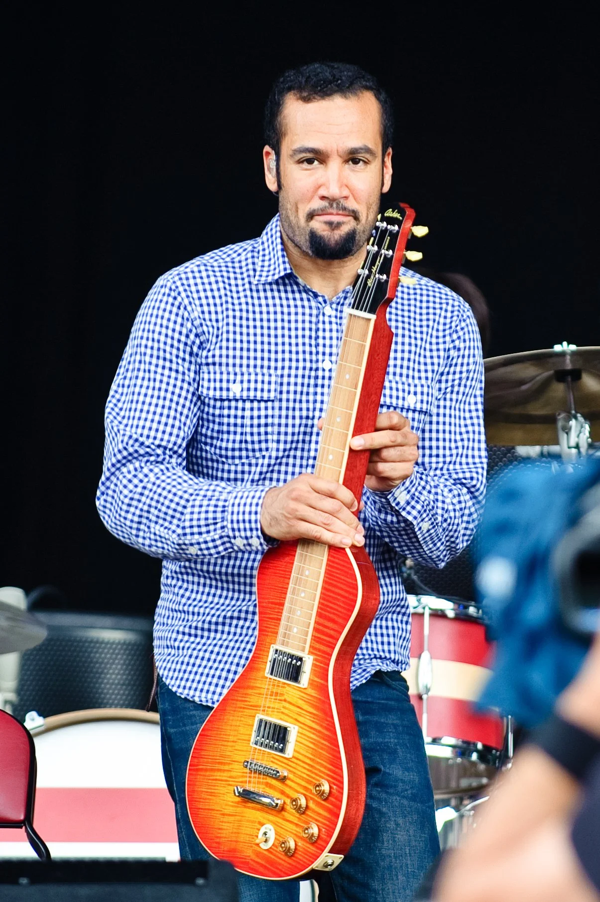 A man with a beard in a blue checkered shirt holding a red electric guitar, standing in front of a drum set on a black background.