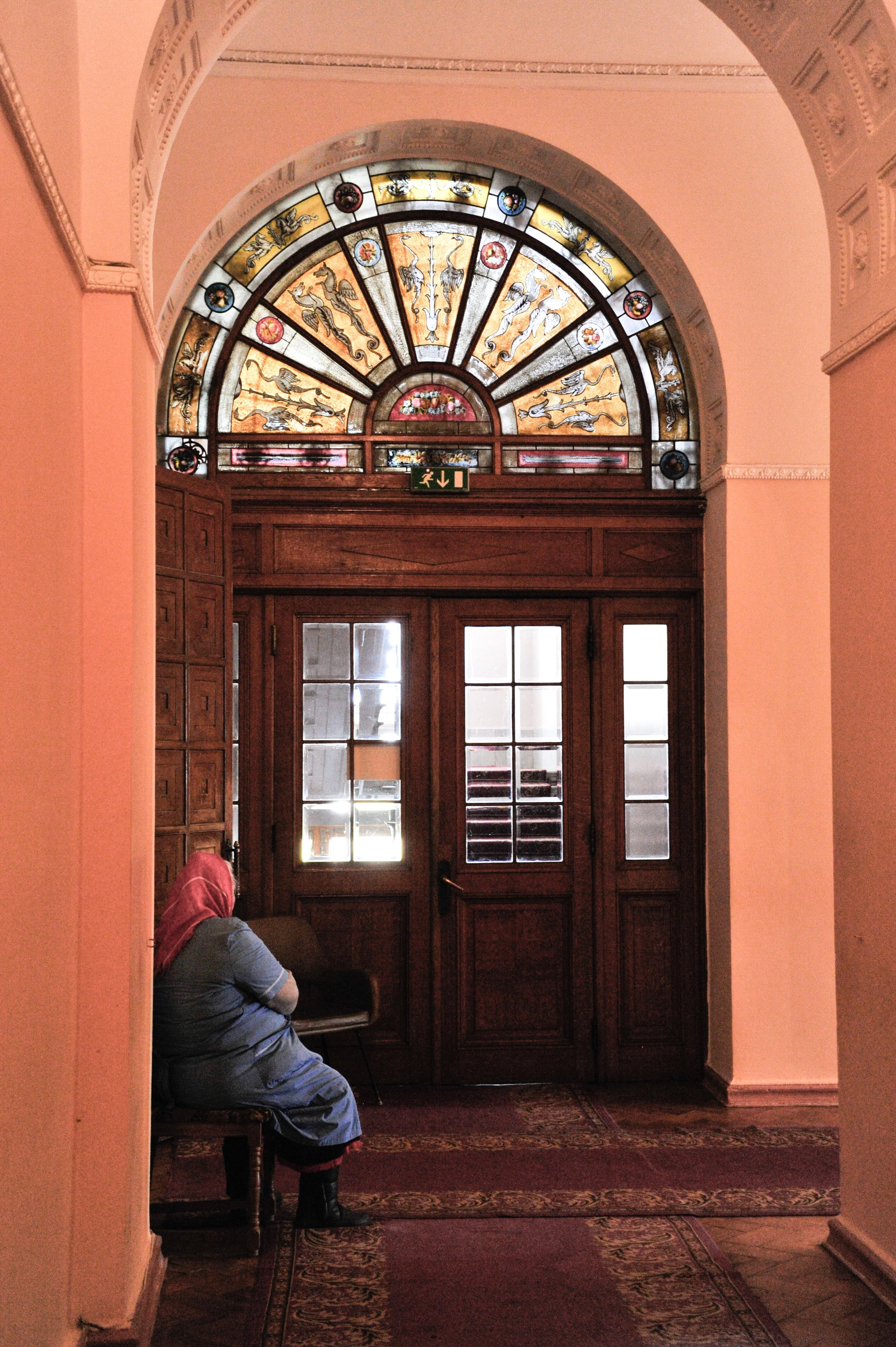 A woman wearing a headscarf sitting on a chair in a hallway with a stained glass window above a wooden door.