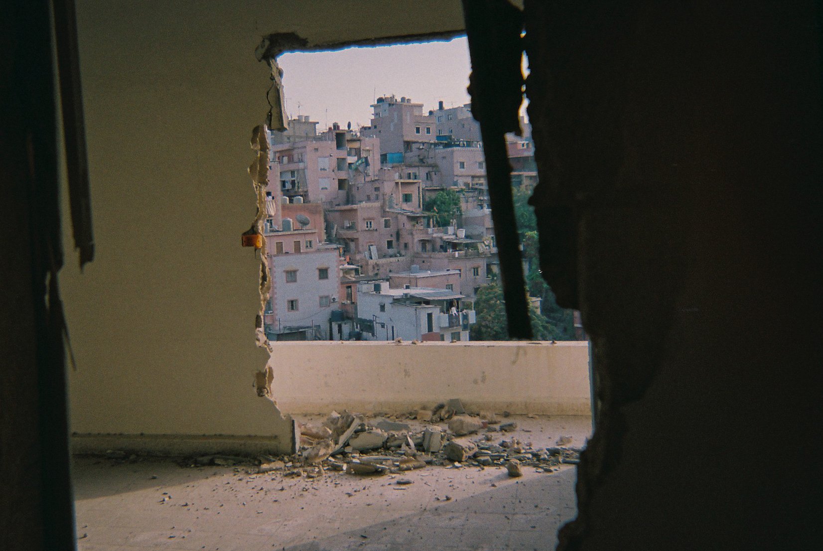 View through a broken wall of a densely populated cityscape with numerous beige and pink buildings on a hillside.
