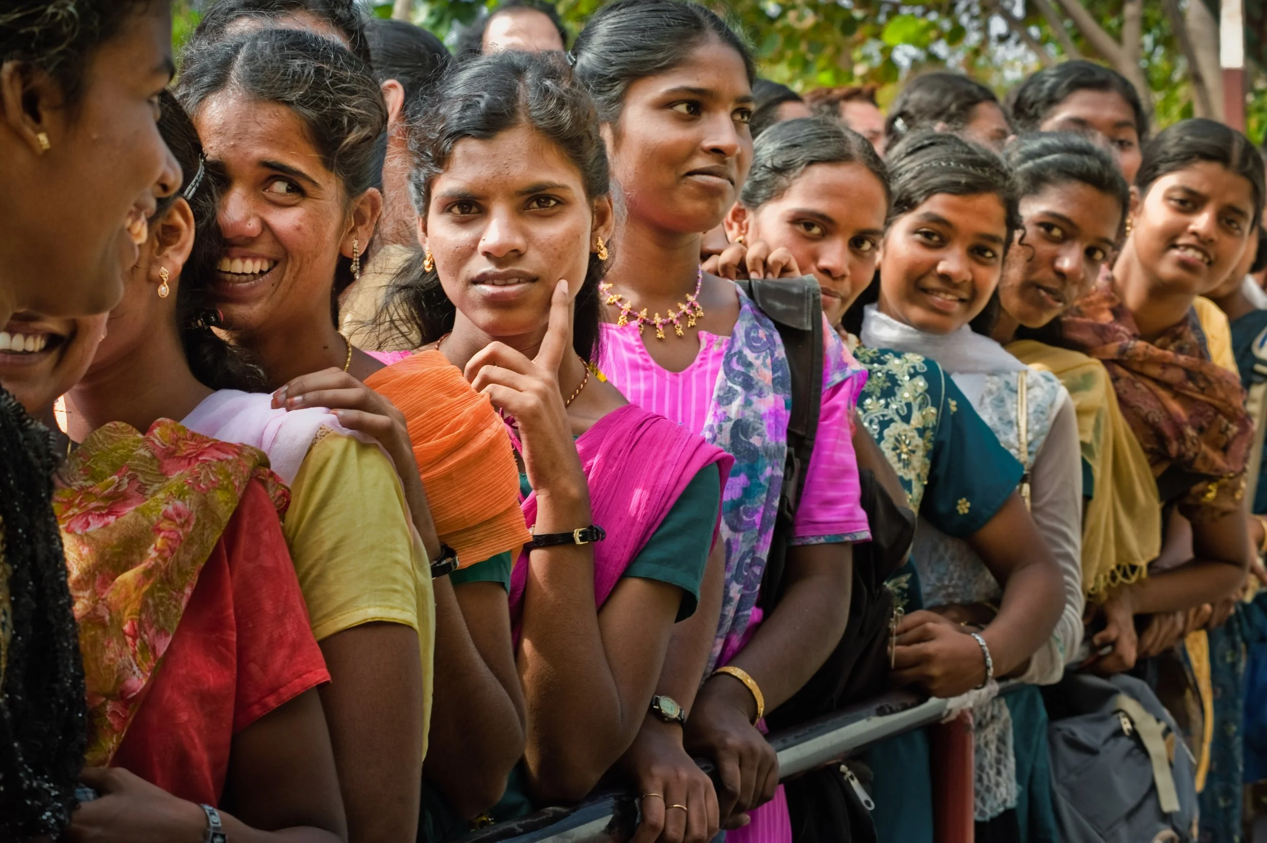 A group of women and girls standing outdoors behind a barrier, dressed in colorful traditional Indian attire, some smiling and some with thoughtful expressions.