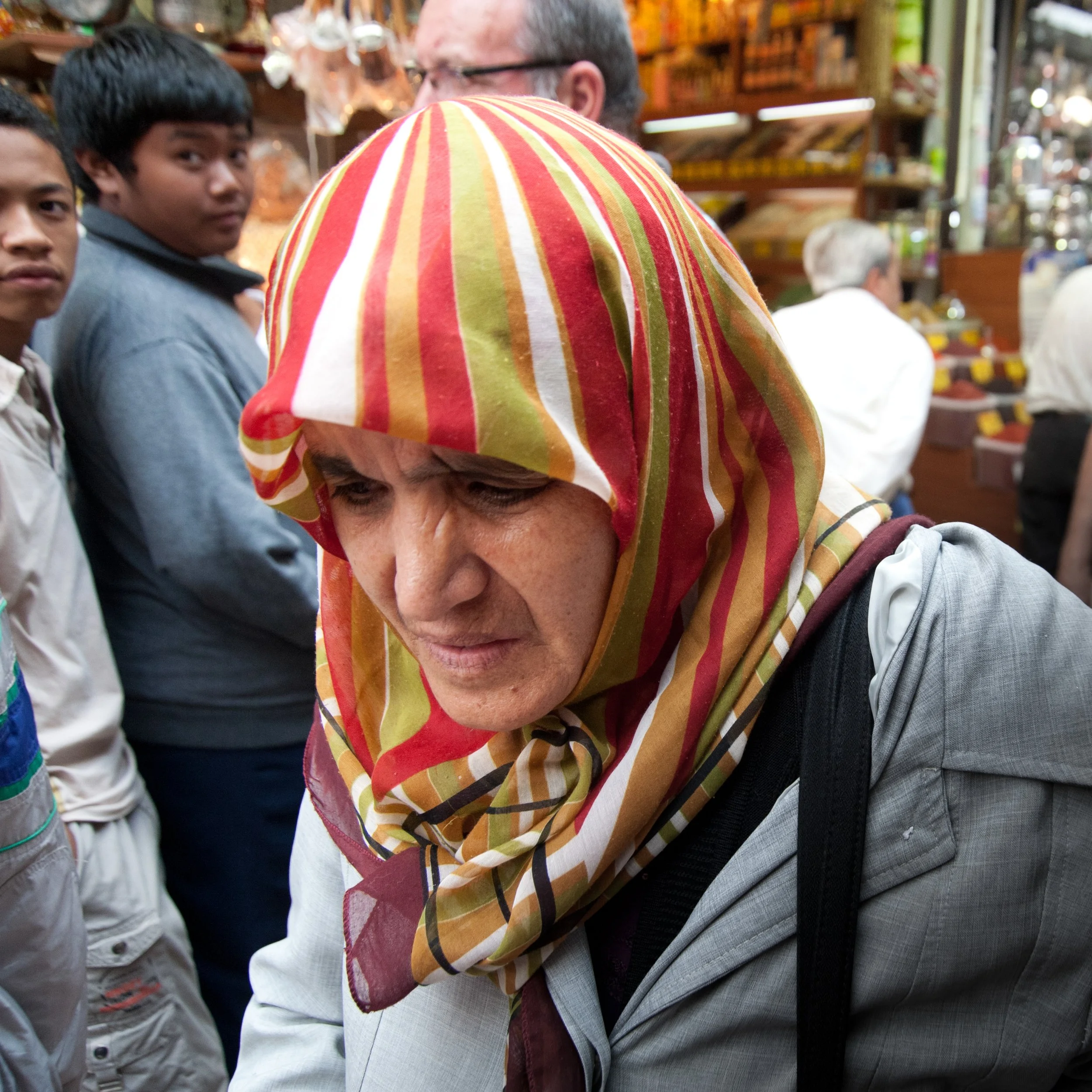 A woman wearing a colorful striped headscarf looking down in a busy marketplace with other people and wooden shelves in the background.