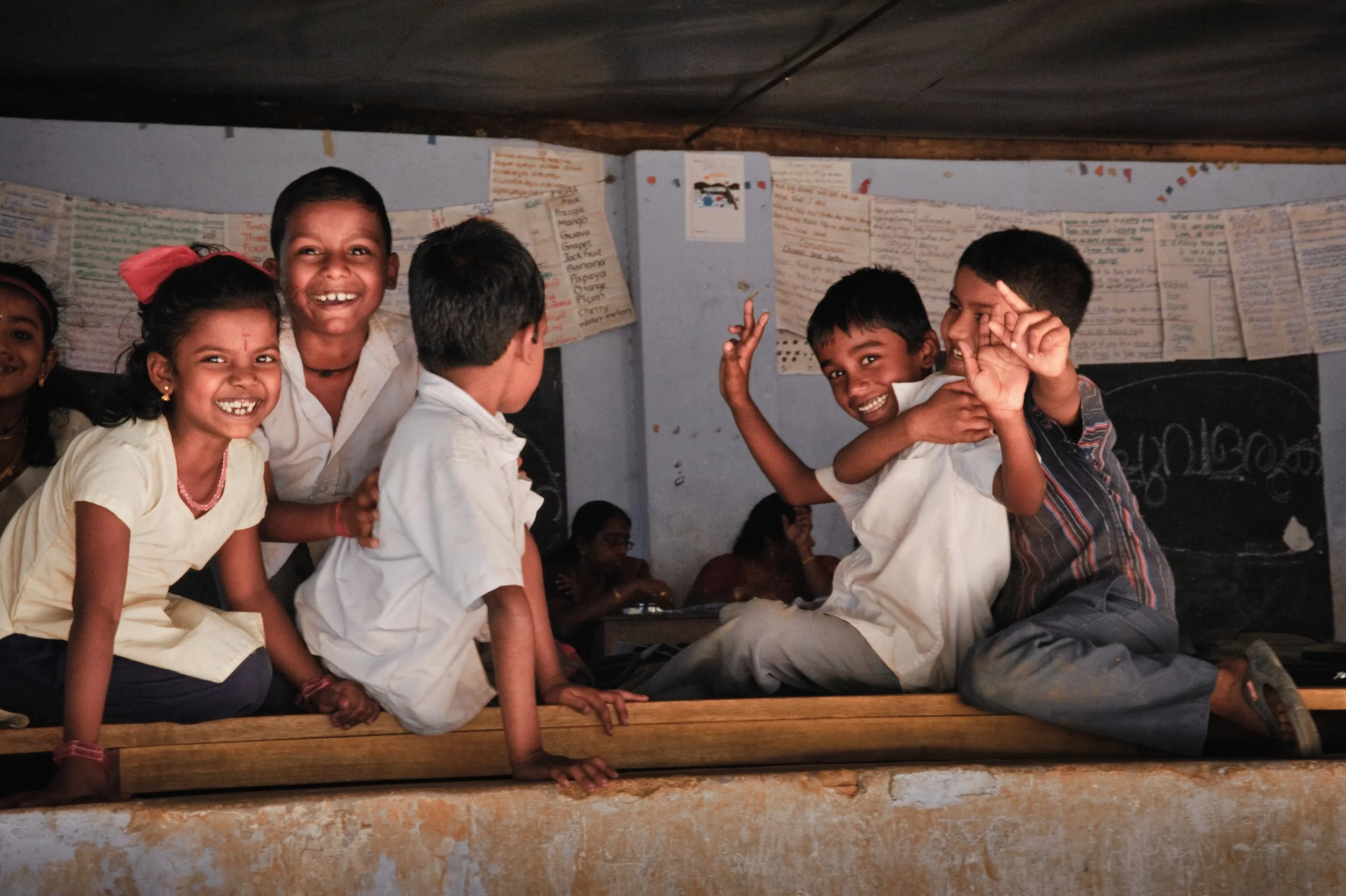Group of smiling children sitting on a wooden platform inside a classroom, some children are playing and laughing, with educational posters on the background wall.