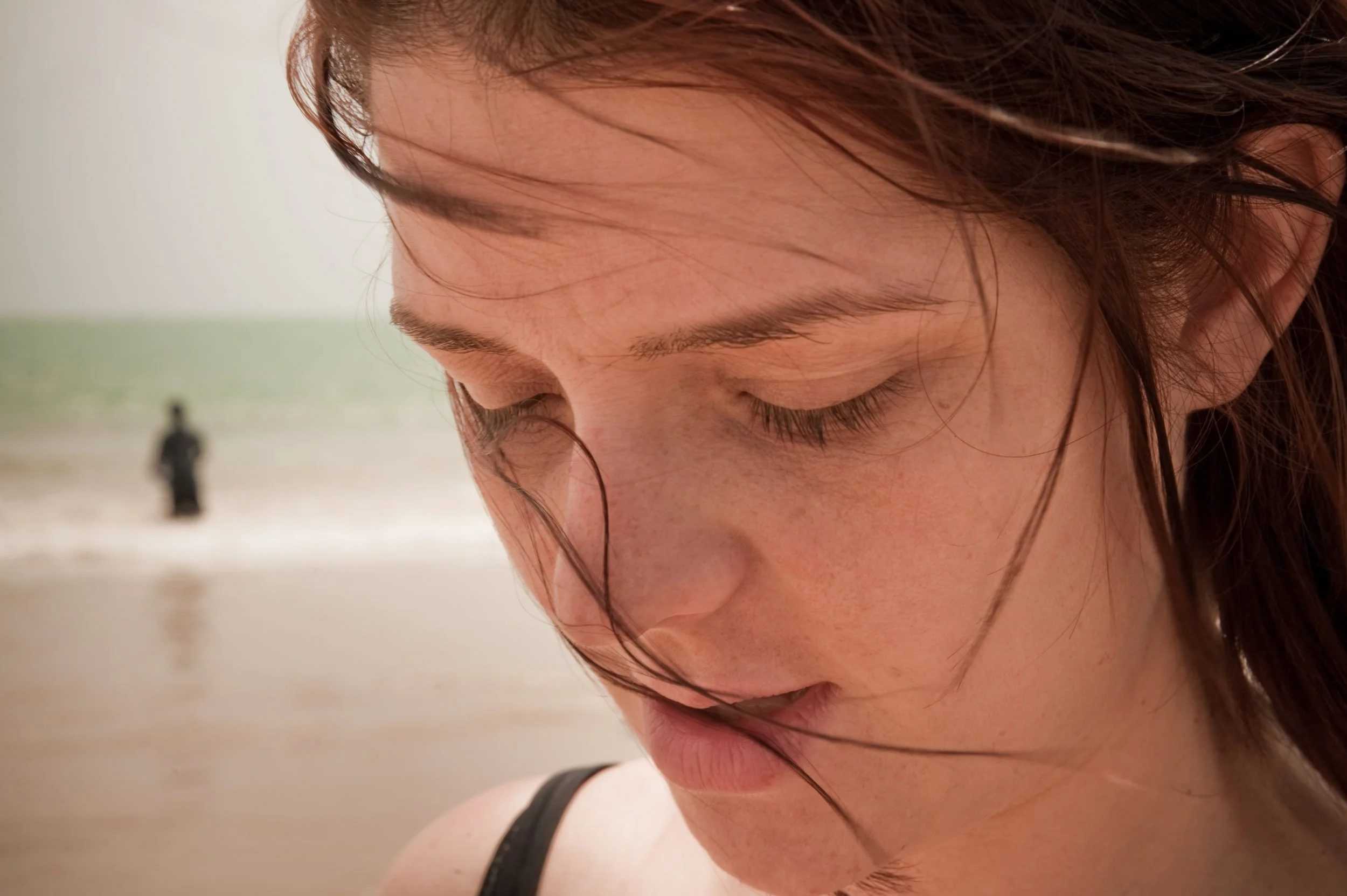 Close-up of a woman's face with closed eyes and wet hair at the beach, with an out-of-focus person and the ocean in the background.