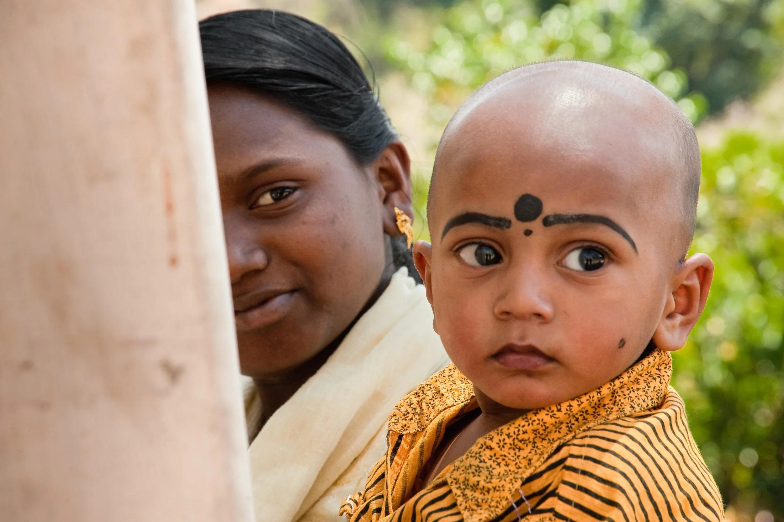 A young boy with a shaved head and facial markings, wearing a striped shirt, looking to the side, with a woman partially visible behind a tree, peeking out from behind it.
