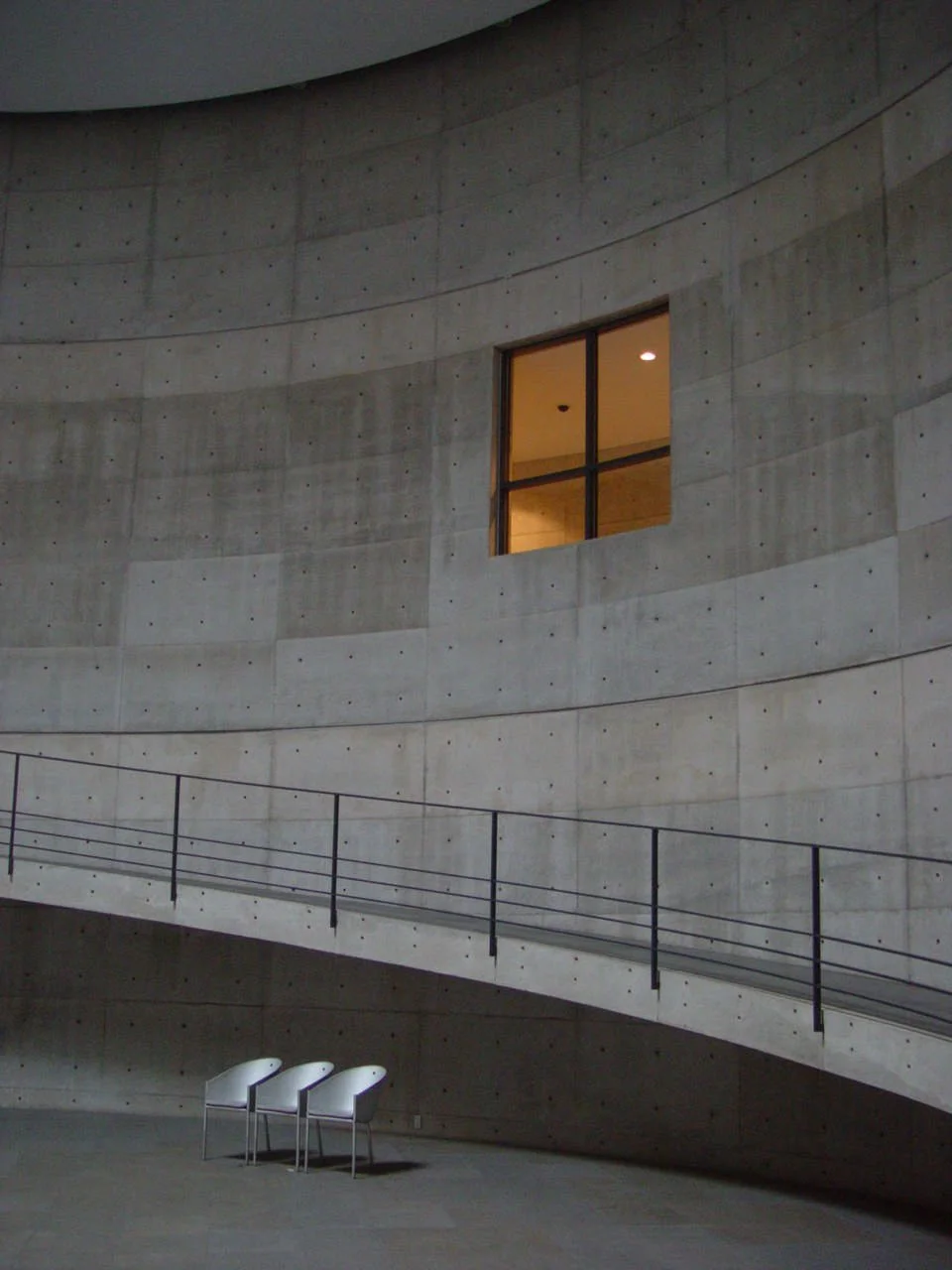 Interior view of a modern concrete building with a curved wall, a single illuminated window, three white chairs, and a walkway with a metal railing.