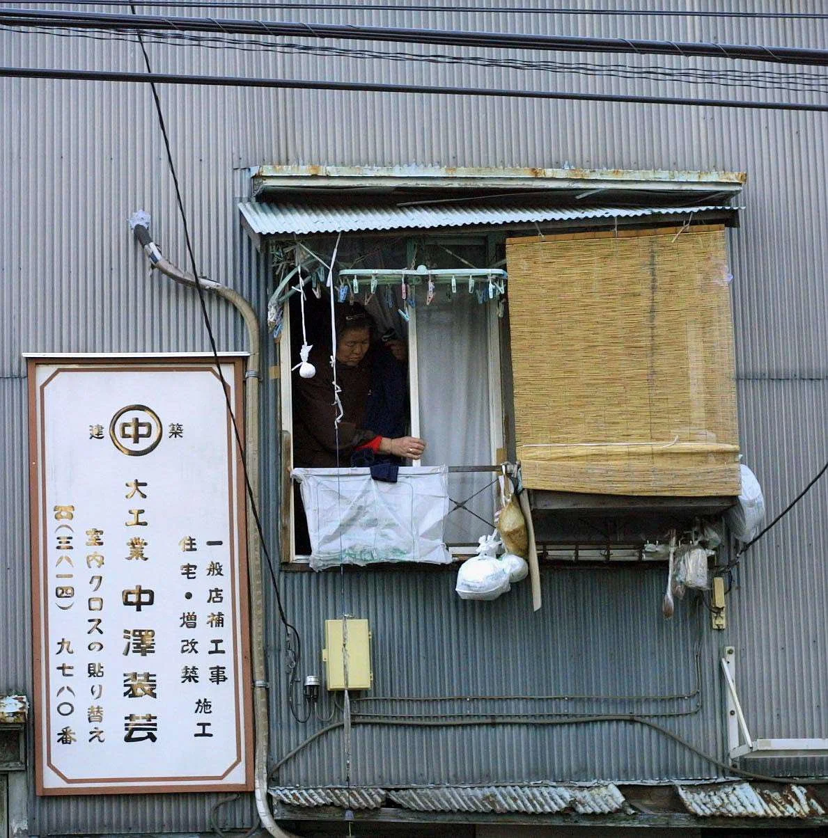 A woman looks out of a small window on a gray metal building with a sign in Japanese to the left and a cluttered exterior including a bamboo blind and various hanging items.