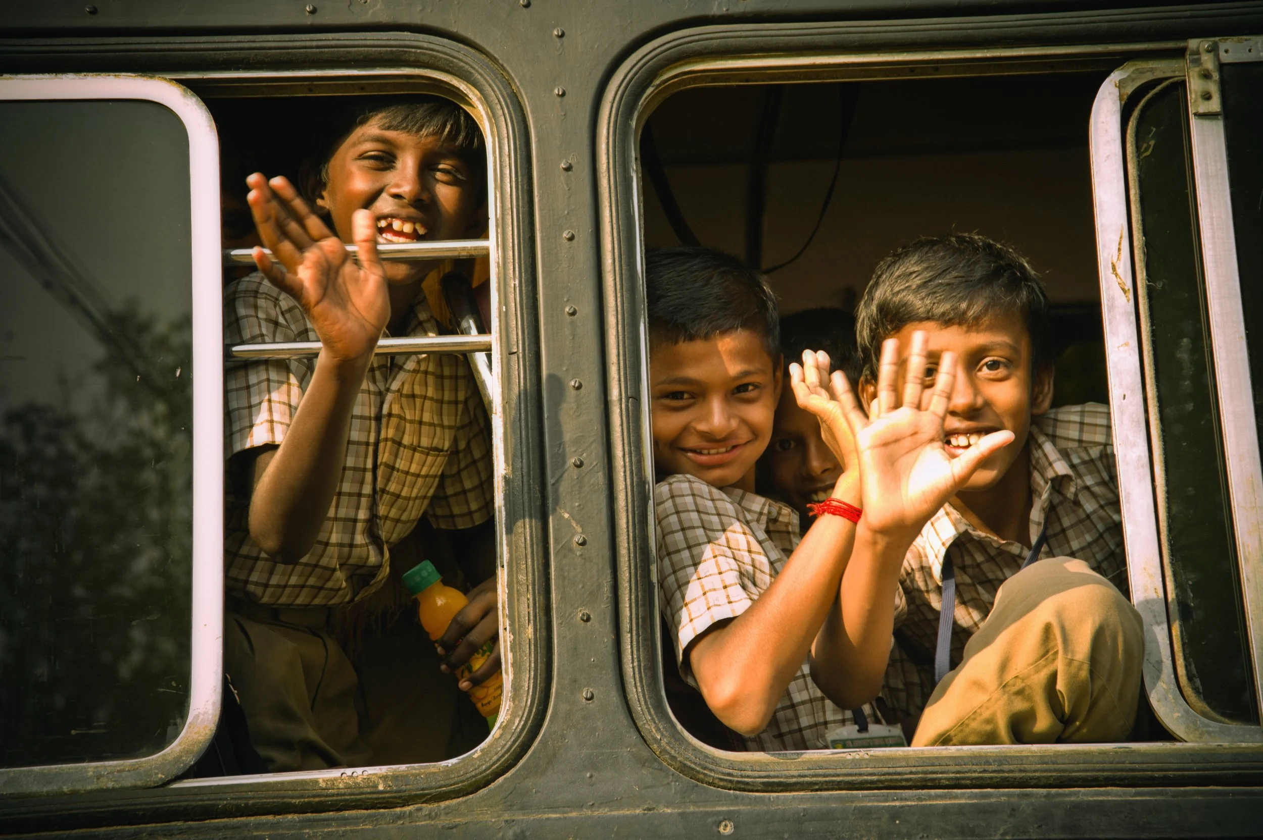 Group of children in school uniforms smiling and waving from windows of a vehicle, possibly a school bus.