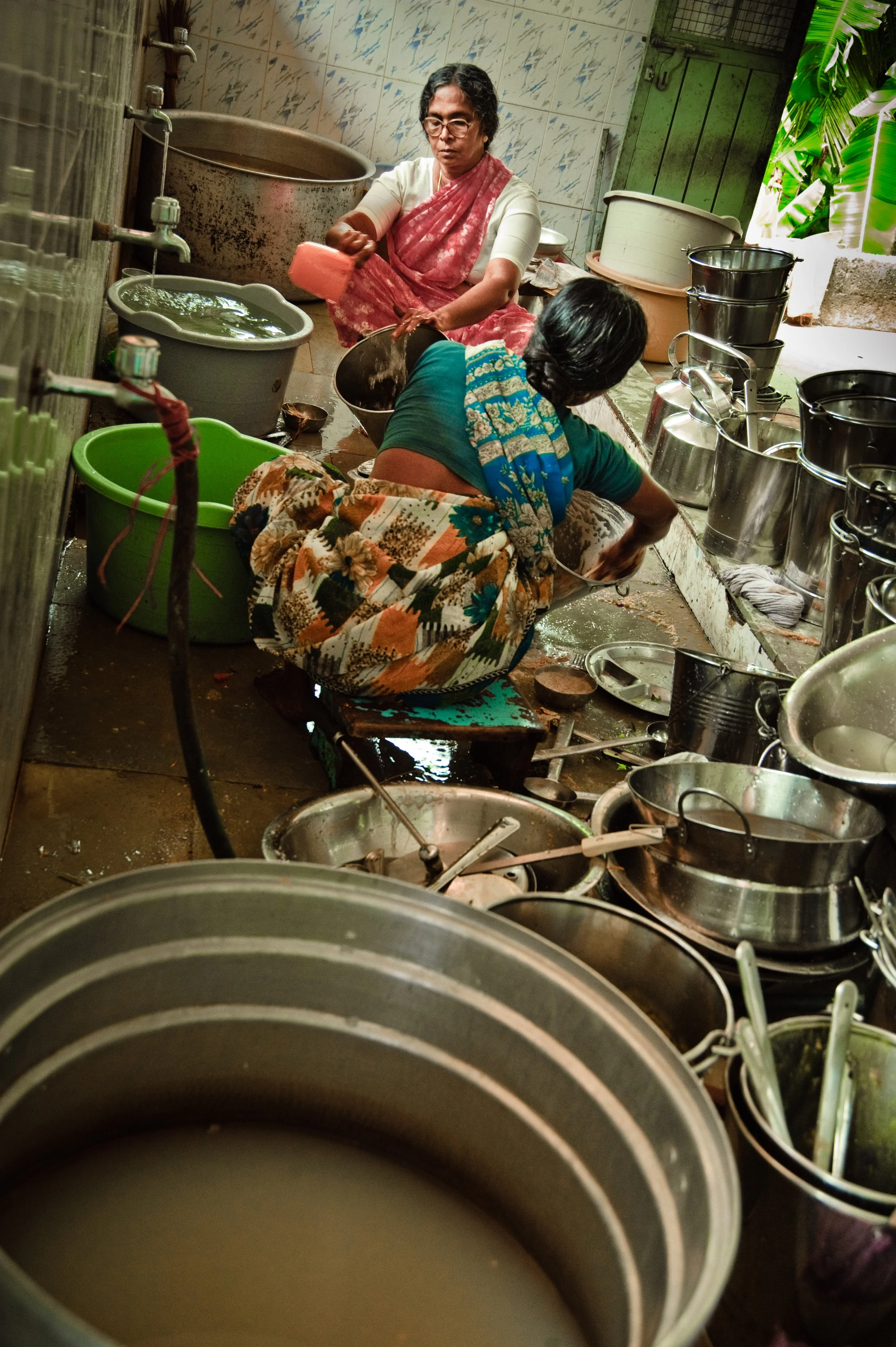 Two women working in a small, crowded kitchen with various pots and utensils, preparing or cooking food, with tiled walls and greenery outside.