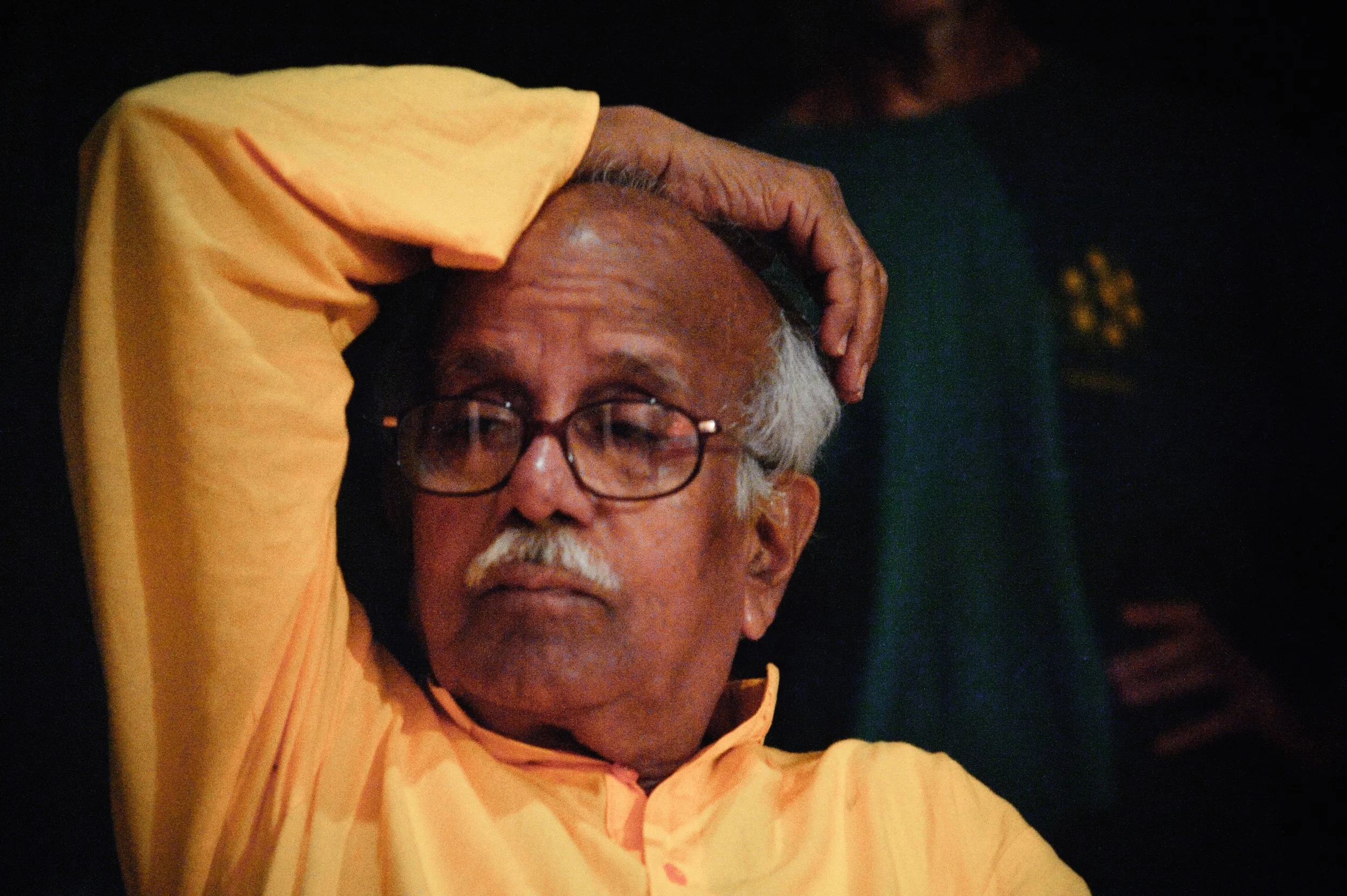 An elderly man with gray hair, glasses, and a mustache wearing a yellow shirt, resting his hand on his head.