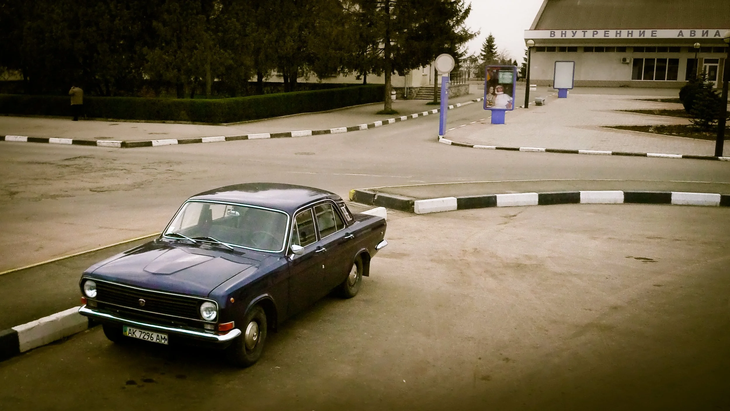 A vintage black car parked at a gas station in a quiet area, with trees and a building in the background.