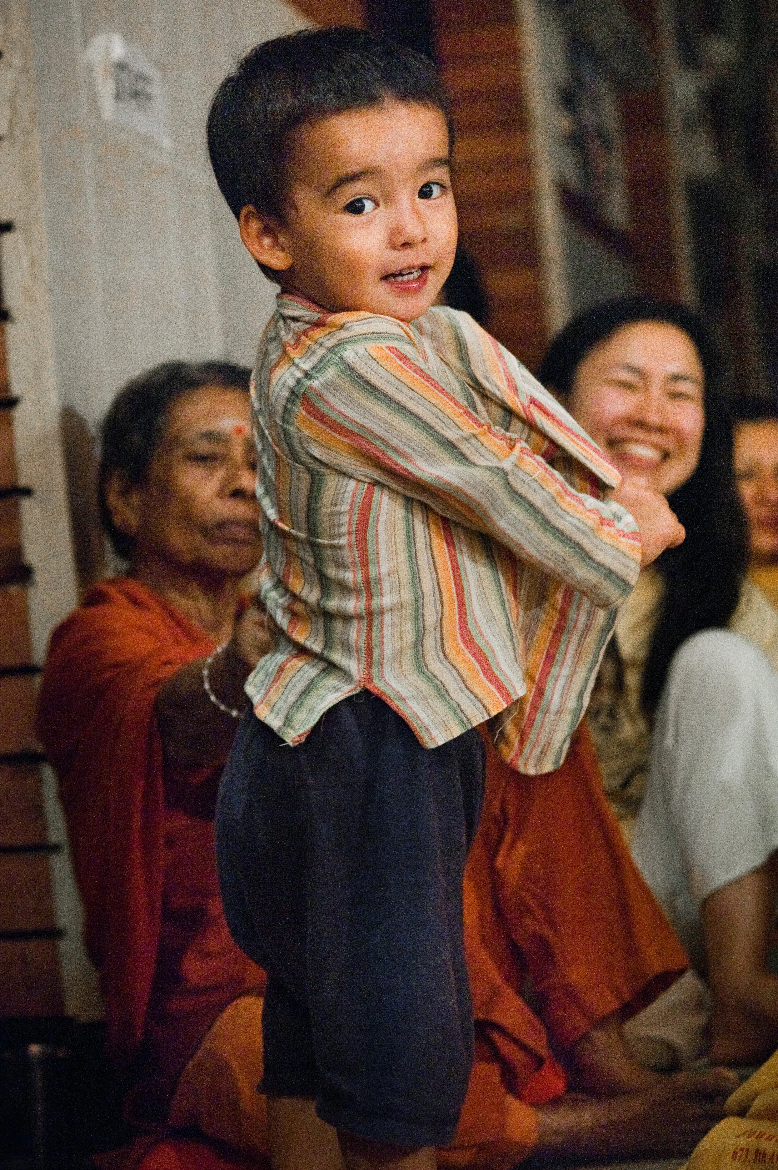 A young boy wearing a striped shirt and dark shorts is smiling and holding onto an adult. Behind him, there are three women, one dressed in orange with a red bindi on her forehead, and they are in a room with wooden walls.