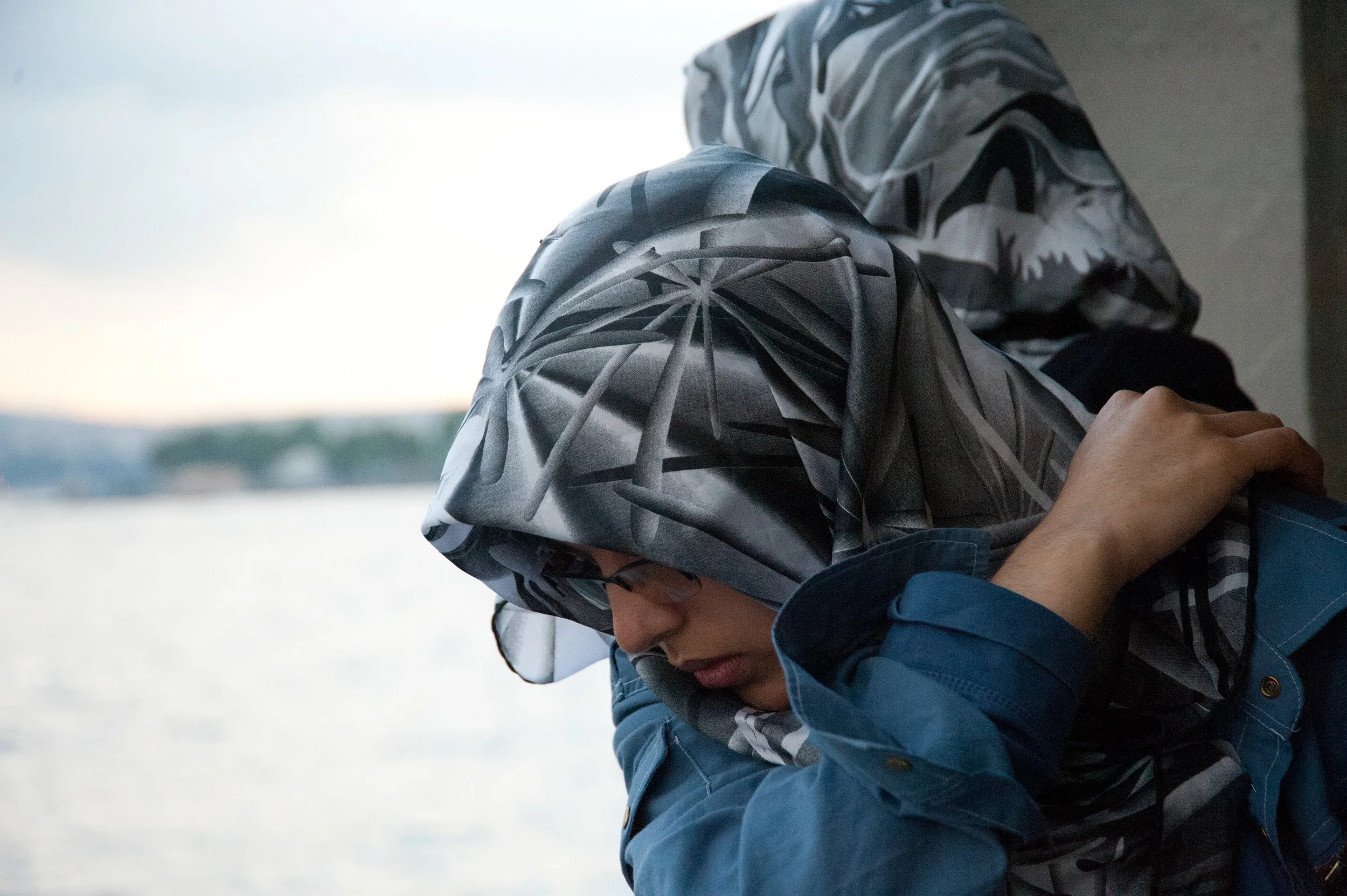Person wearing glasses and a gray geometric patterned headscarf, looking down, by a body of water during daytime.