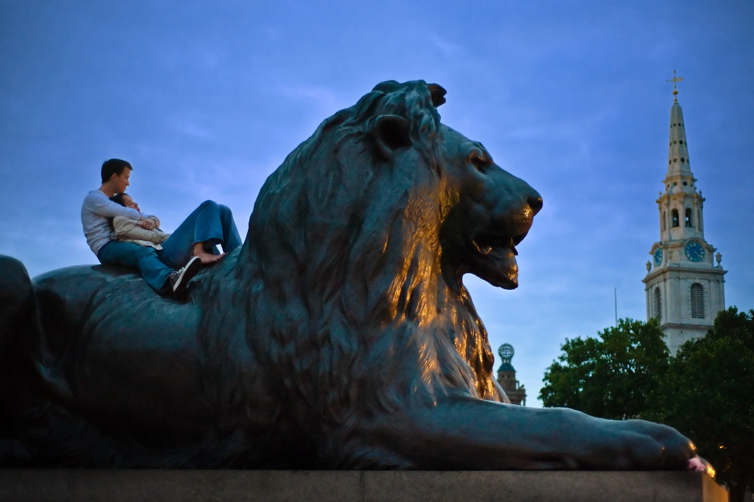 Two people sitting on a large bronze lion statue in a park during dusk, with a historic church steeple and trees in the background.