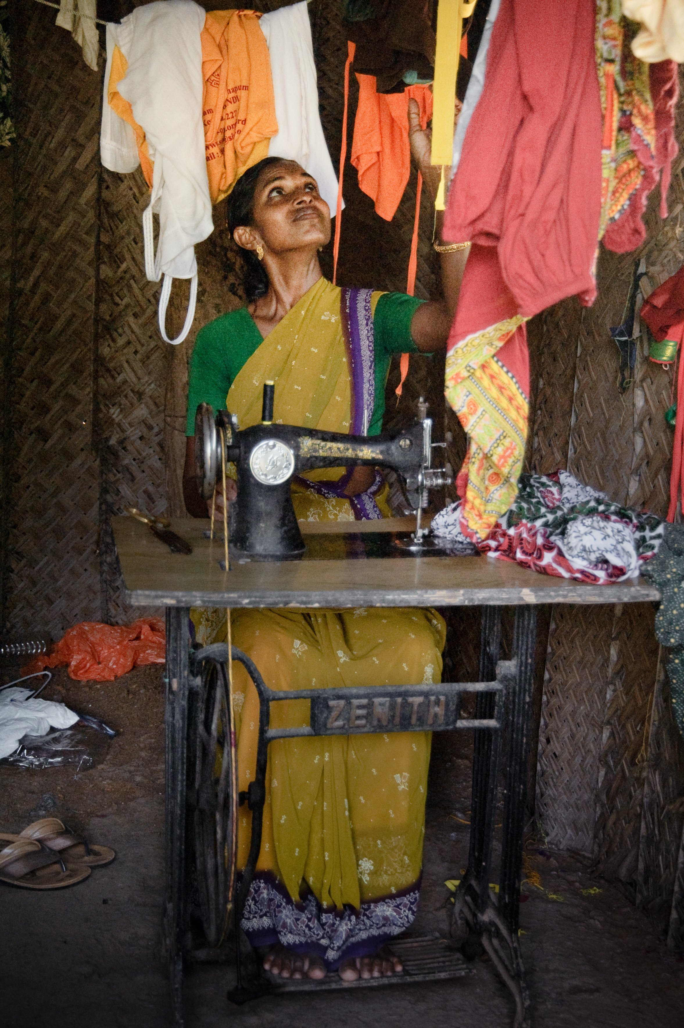 A woman sitting at a sewing machine in a small, bamboo-walled workspace, sewing colorful fabric. She is wearing a green blouse and a yellow sari with purple border, surrounded by hanging clothes and a cluttered environment.