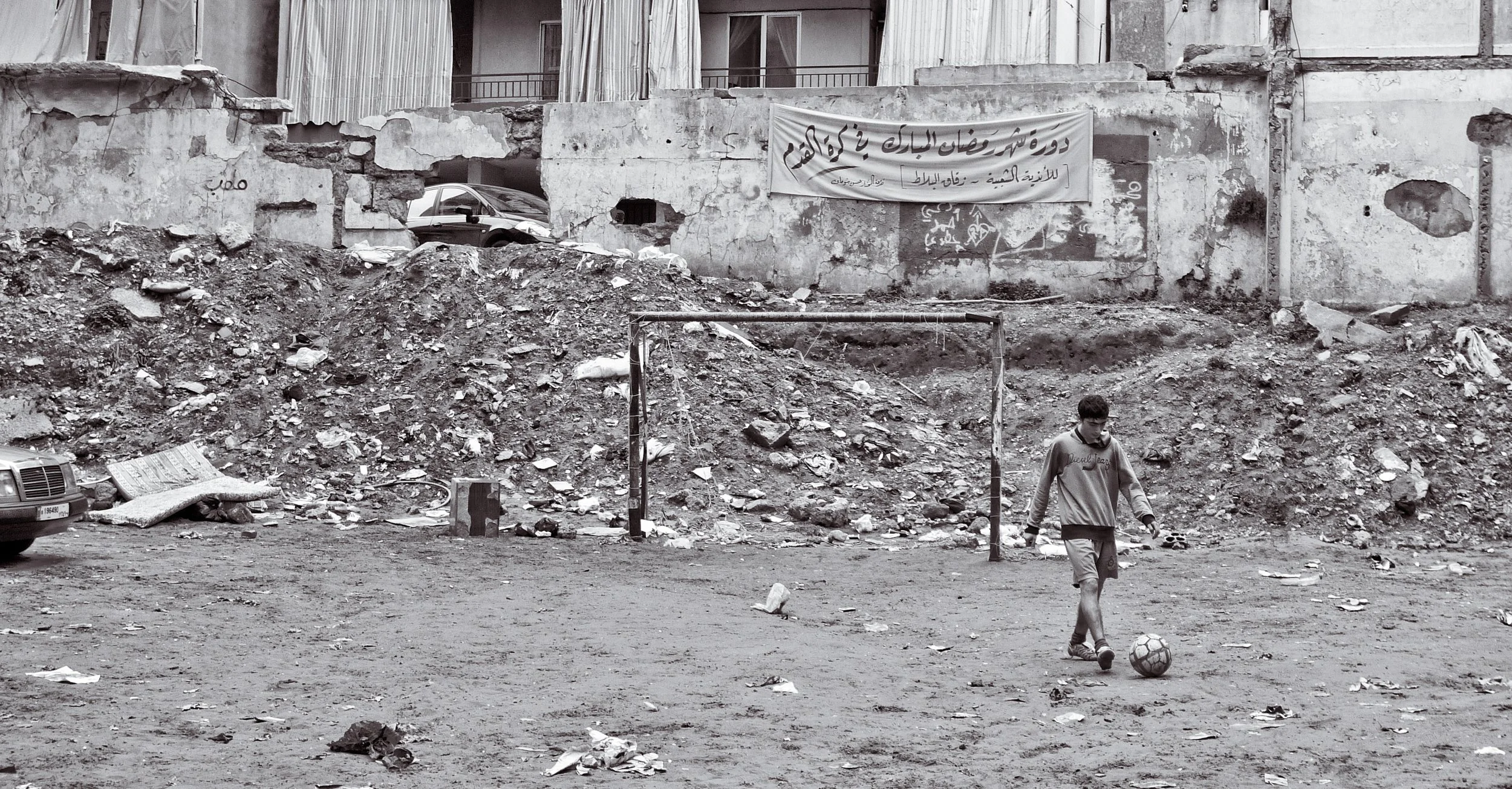A young person playing soccer on a dirt field in front of a ruined concrete wall with debris and a banner in Arabic.