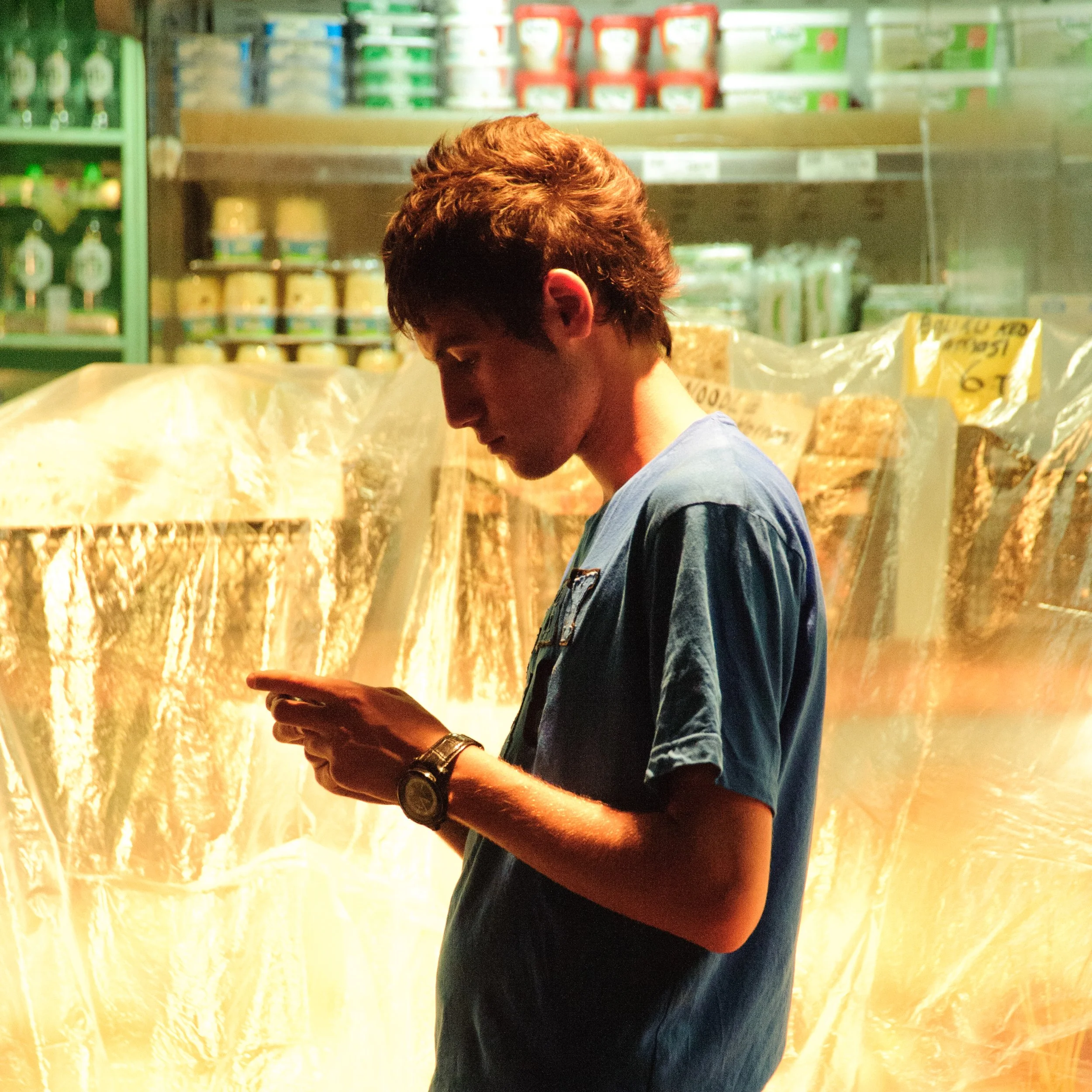 A young man in a blue shirt standing in a grocery store aisle, looking at his phone, with shelves of food products behind him.