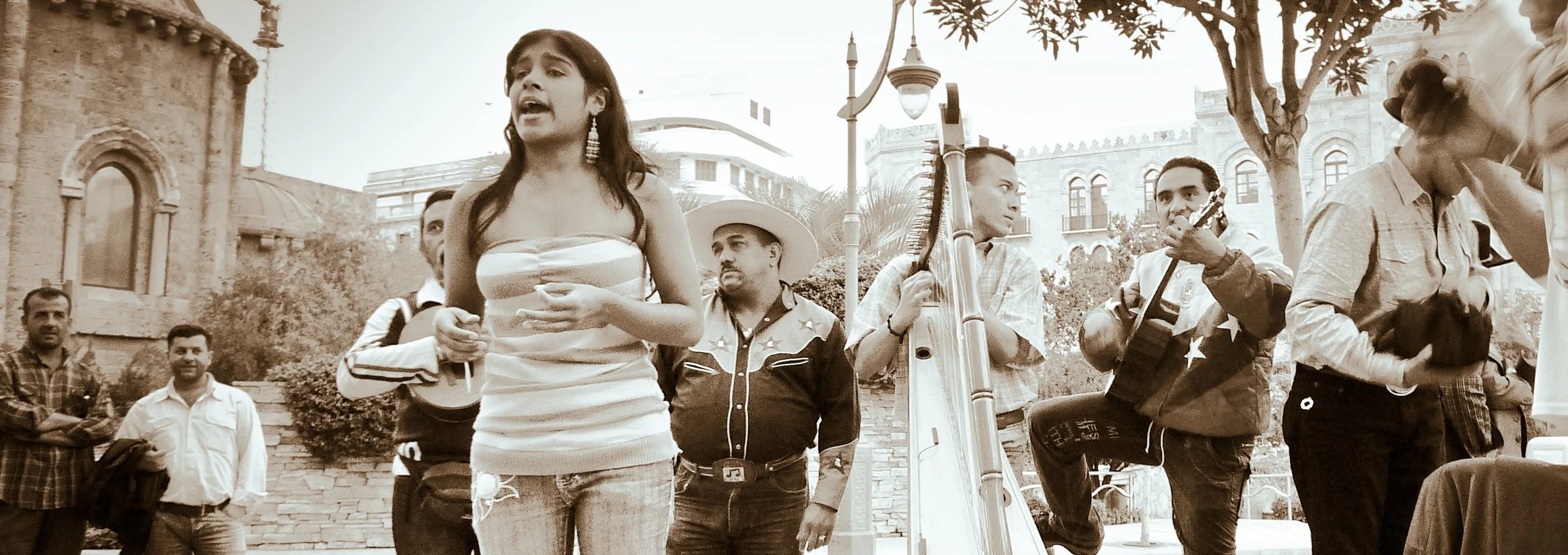 A group of people gathered outdoors, some playing musical instruments and singing, with historic buildings and trees in the background, in a sepia tone.