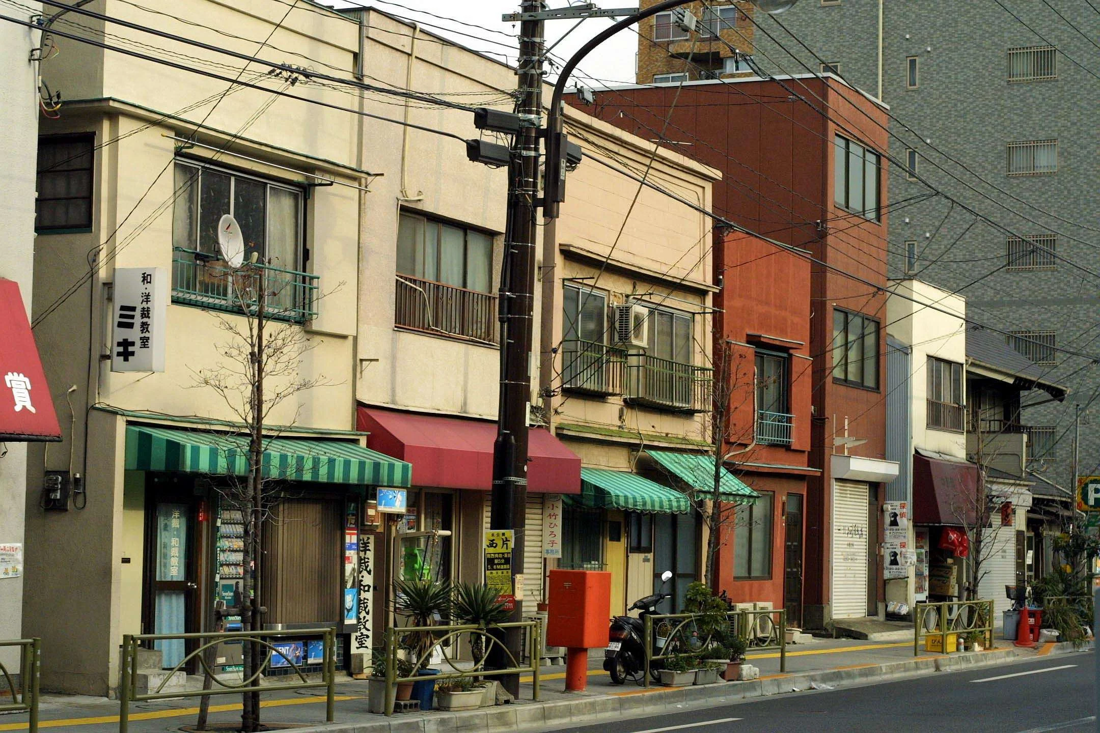 Street view in Japan with small shops, traditional signage, leafless trees, and overhead power lines.
