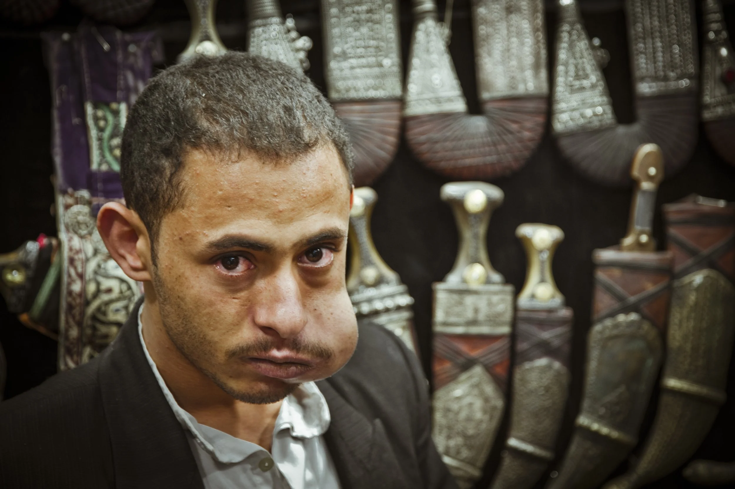 A man with short dark hair and a mustache, wearing a black suit and white shirt, looking at the camera with a serious expression. Behind him are traditional handbags or woven items displayed on a wall.