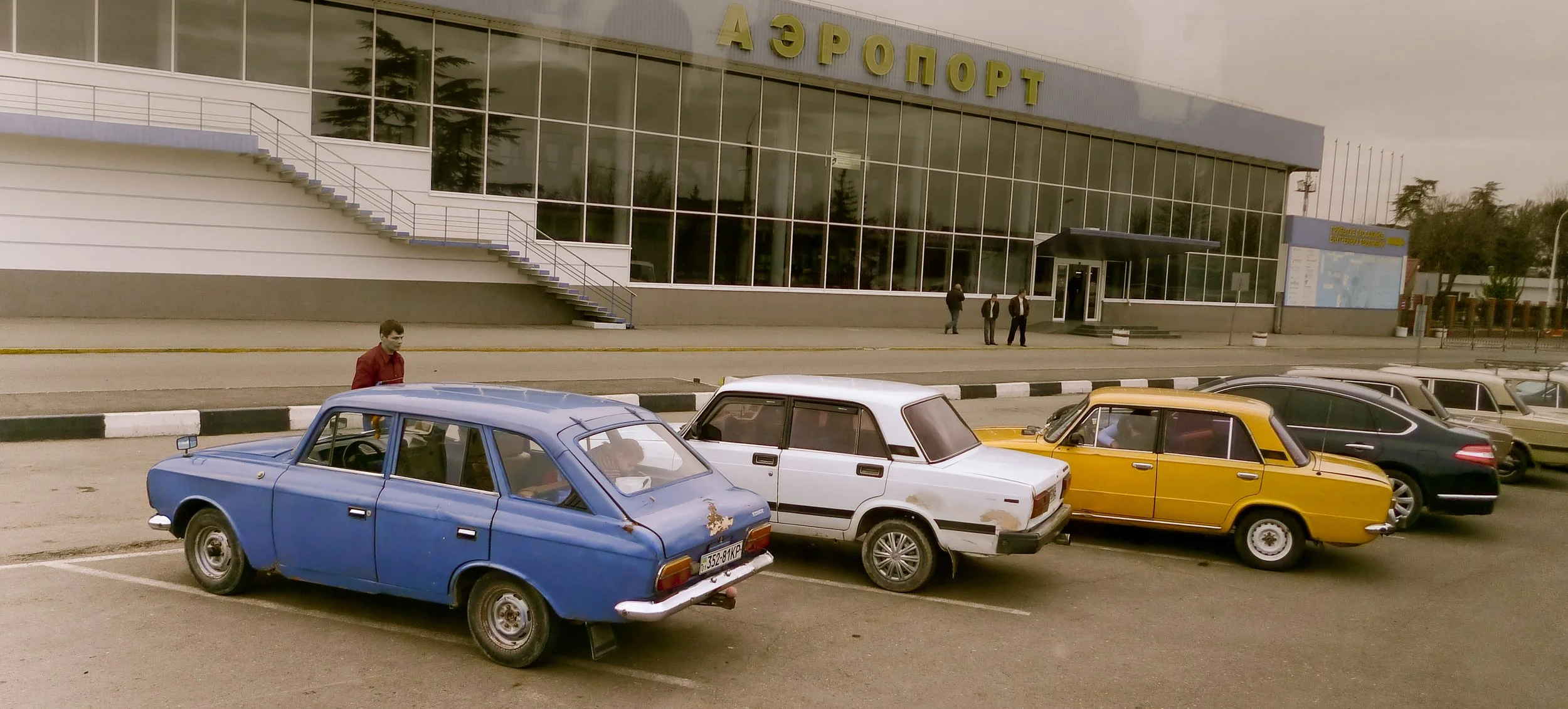 A parking lot in front of a modern glass building with a yellow sign in Russian that says 'Аэропорт' (Airport). Several parked cars, including a blue hatchback, a white sedan, a yellow sedan, and others, are visible. A man stands near the blue car, a
